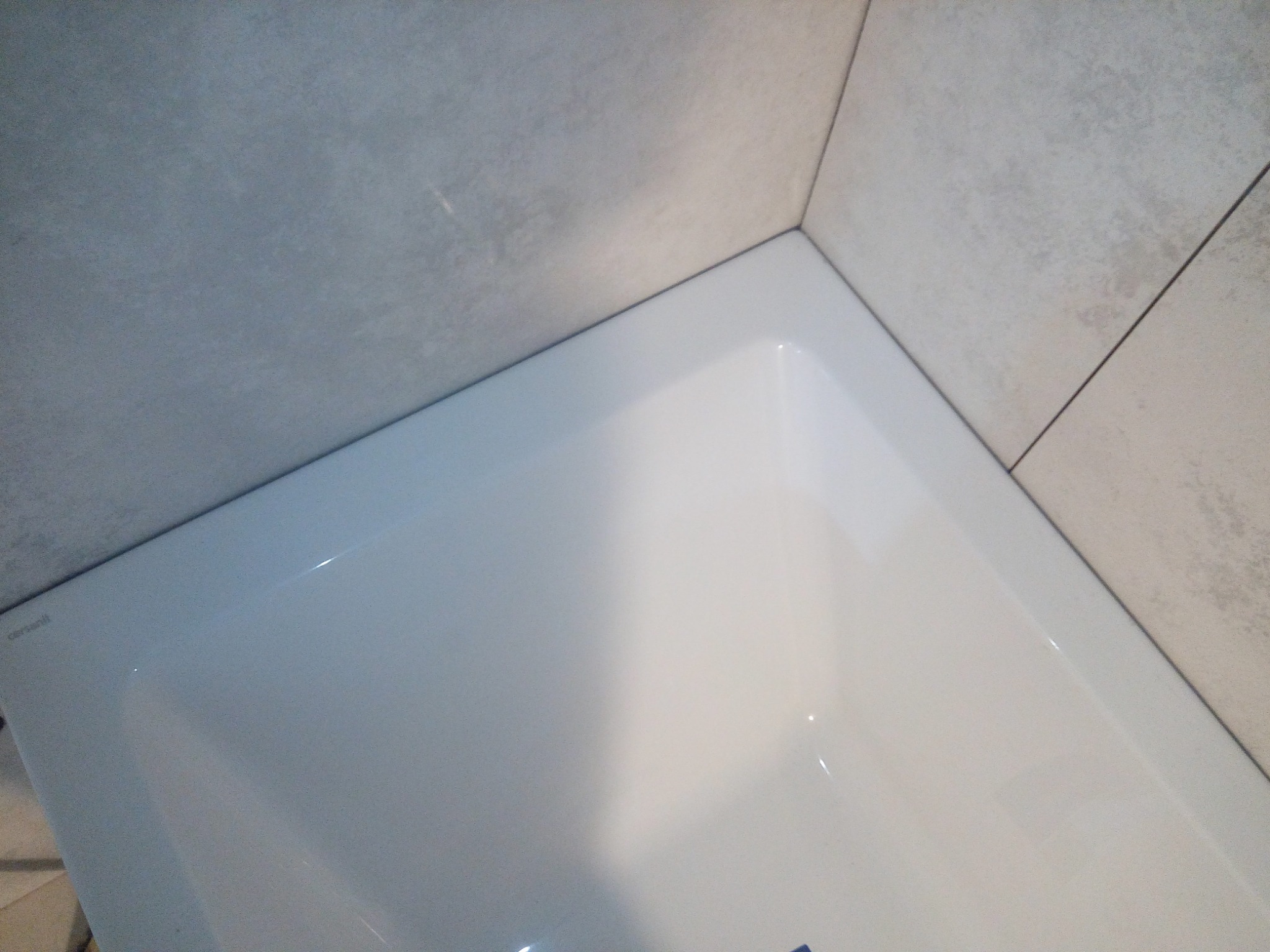 Fragment narożnika łazienki z białą wanną Cersanit i szarymi, matowymi płytkami na ścianach.