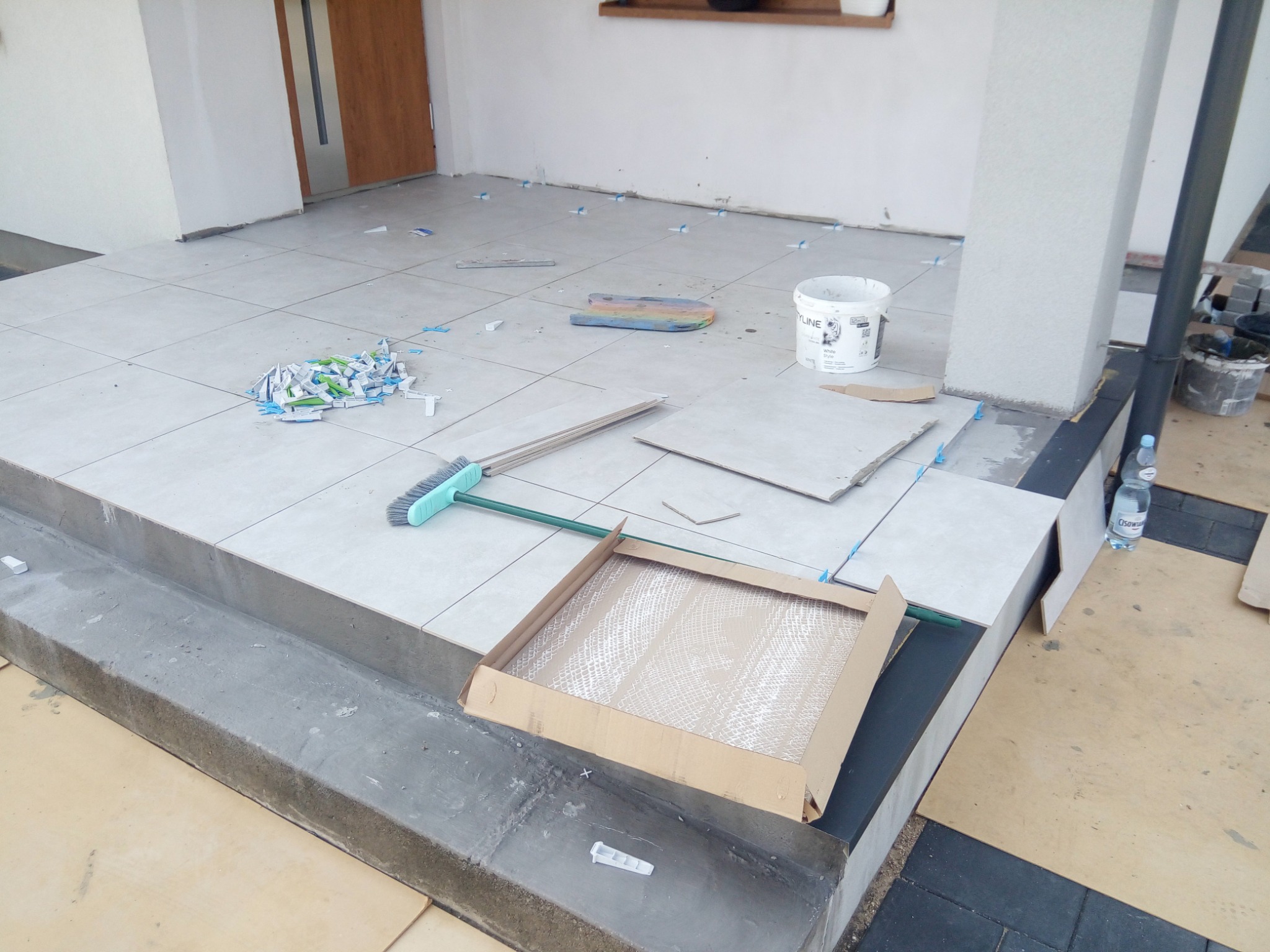 Układanie jasnych płytek podłogowych na zewnątrz budynku, widoczne narzędzia, puste opakowanie po płytkach i pozostałości po elementach dystansowych.