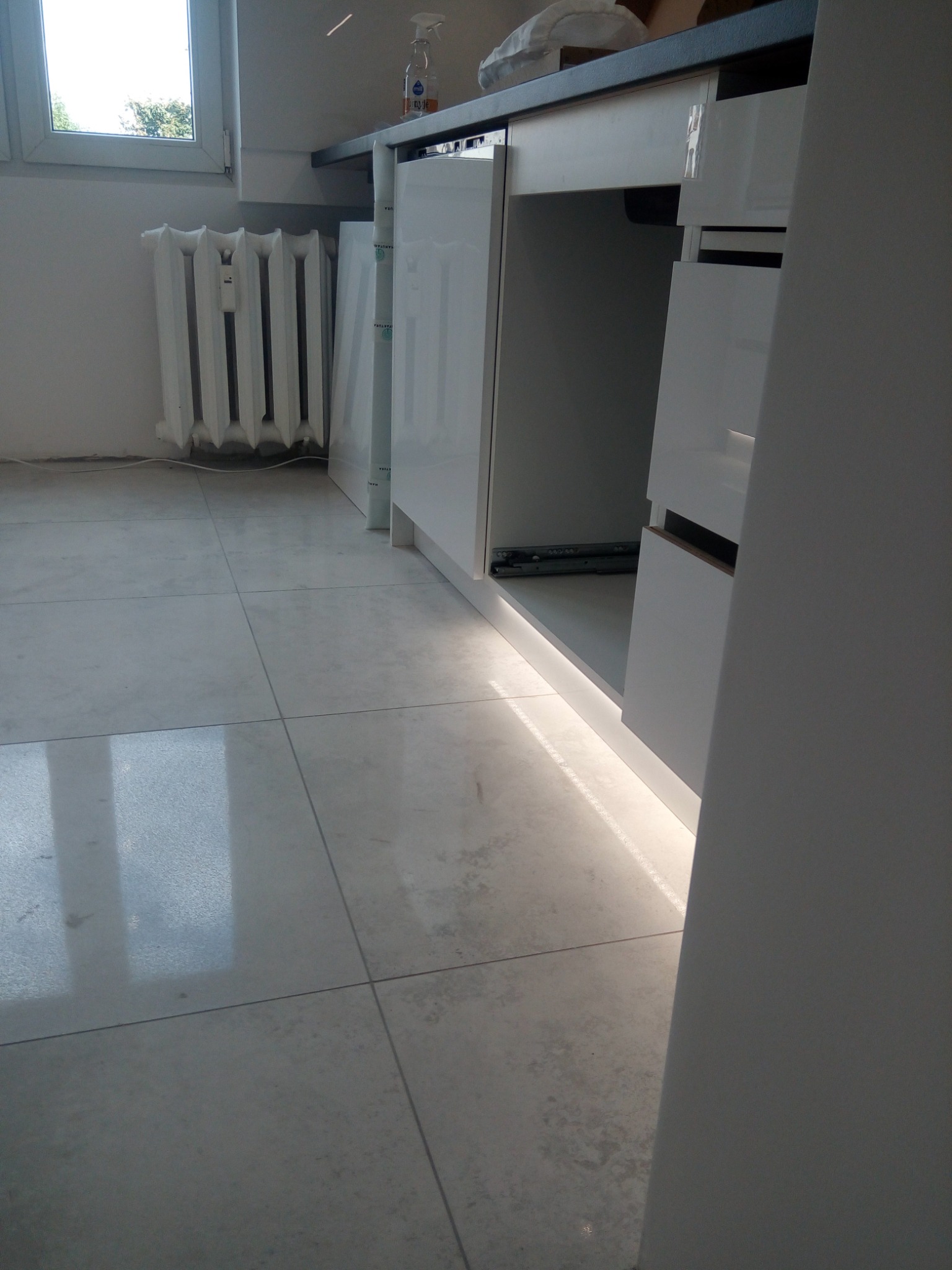 Montaż białych, lakierowanych szafek kuchennych z szufladami, widoczne prowadnice i oświetlenie LED pod szafkami, na podłodze szare płytki, w tle grzejnik.