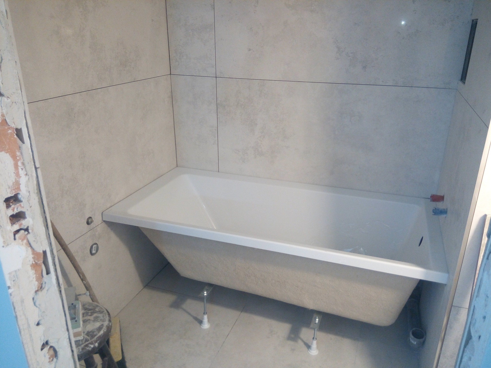 Narożna wanna na regulowanych nóżkach, częściowo obudowana płytkami w odcieniach szarości, widoczne fragmenty niedokończonej ściany i instalacji wodnej.