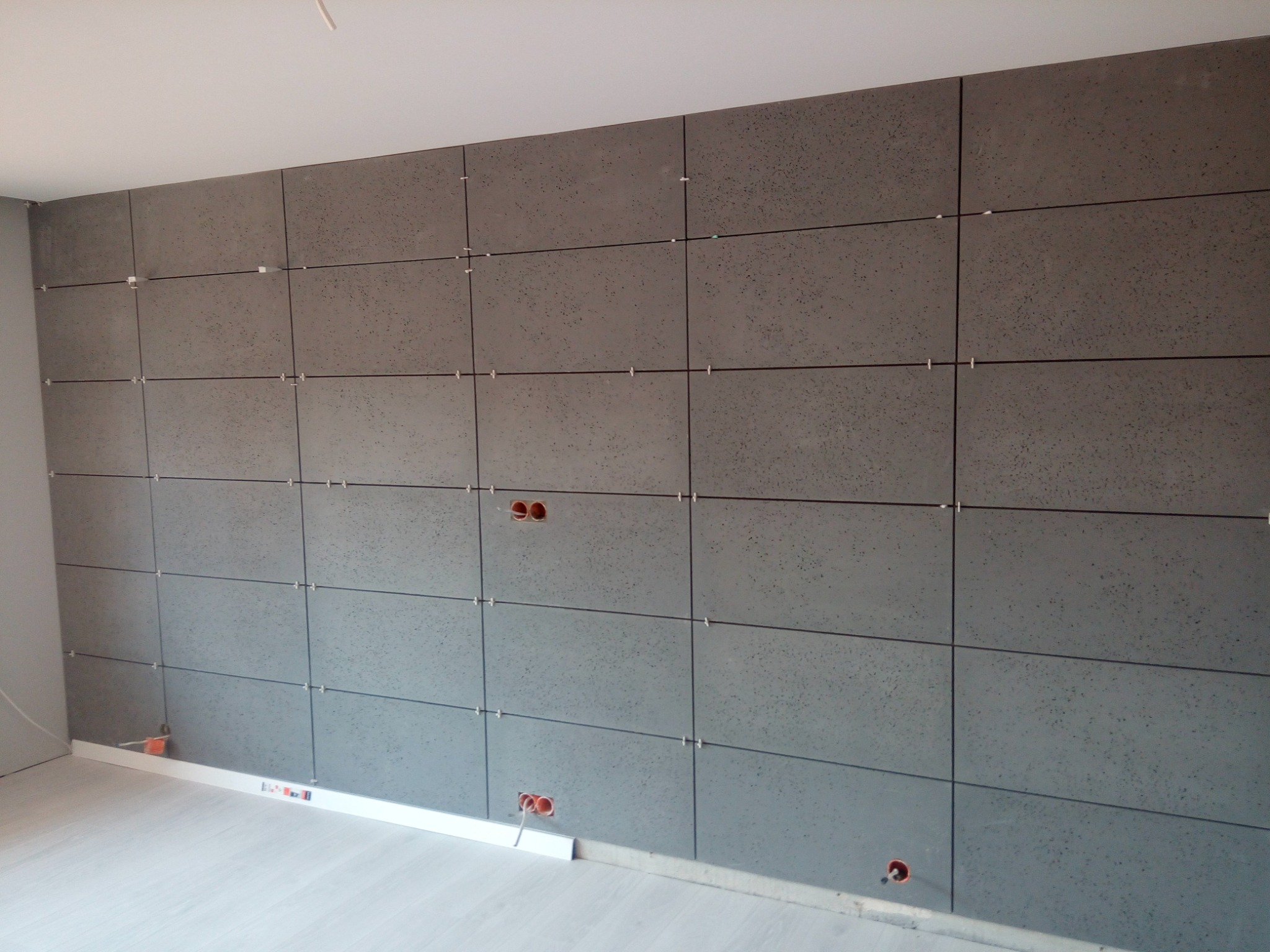 Ściana wyłożona szarymi panelami z widocznymi otworami na gniazdka elektryczne, poziomica przy podłodze i krzyżyki dystansowe między panelami, ukazująca etap prac wykończeniowych.