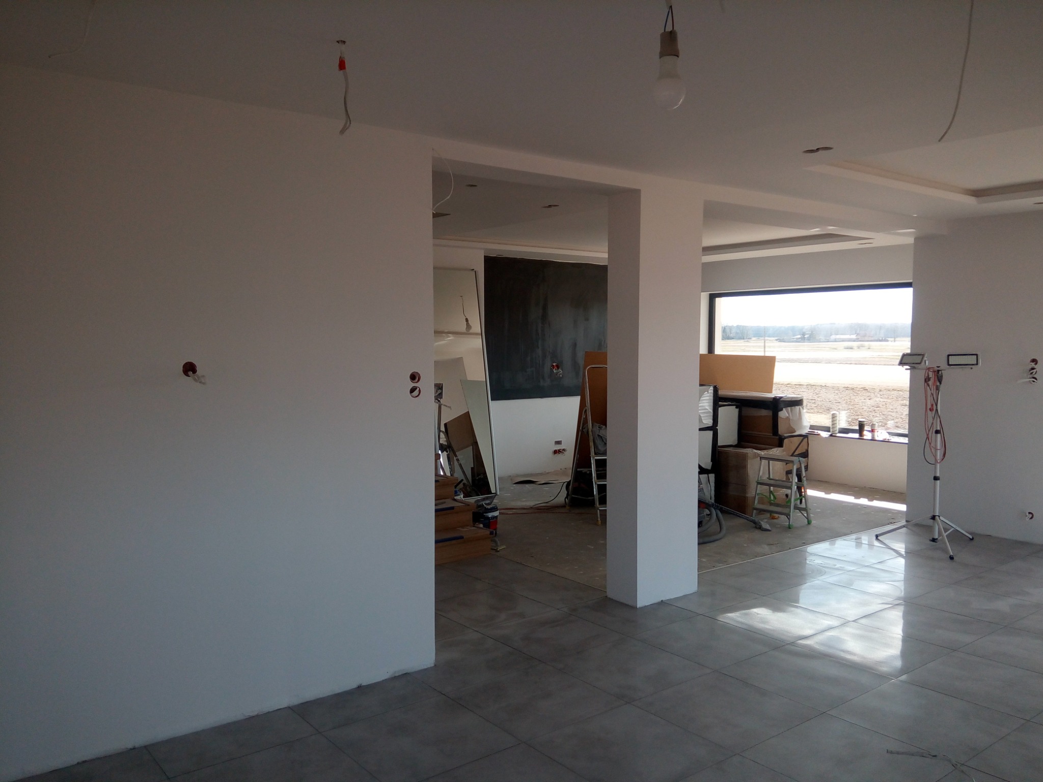 Surowe wnętrze w trakcie prac wykończeniowych: świeżo wylana podłoga z płytek, białe ściany z widocznymi przewodami, sufit podwieszany z oświetleniem, widok na zewnątrz przez okno, drabina...