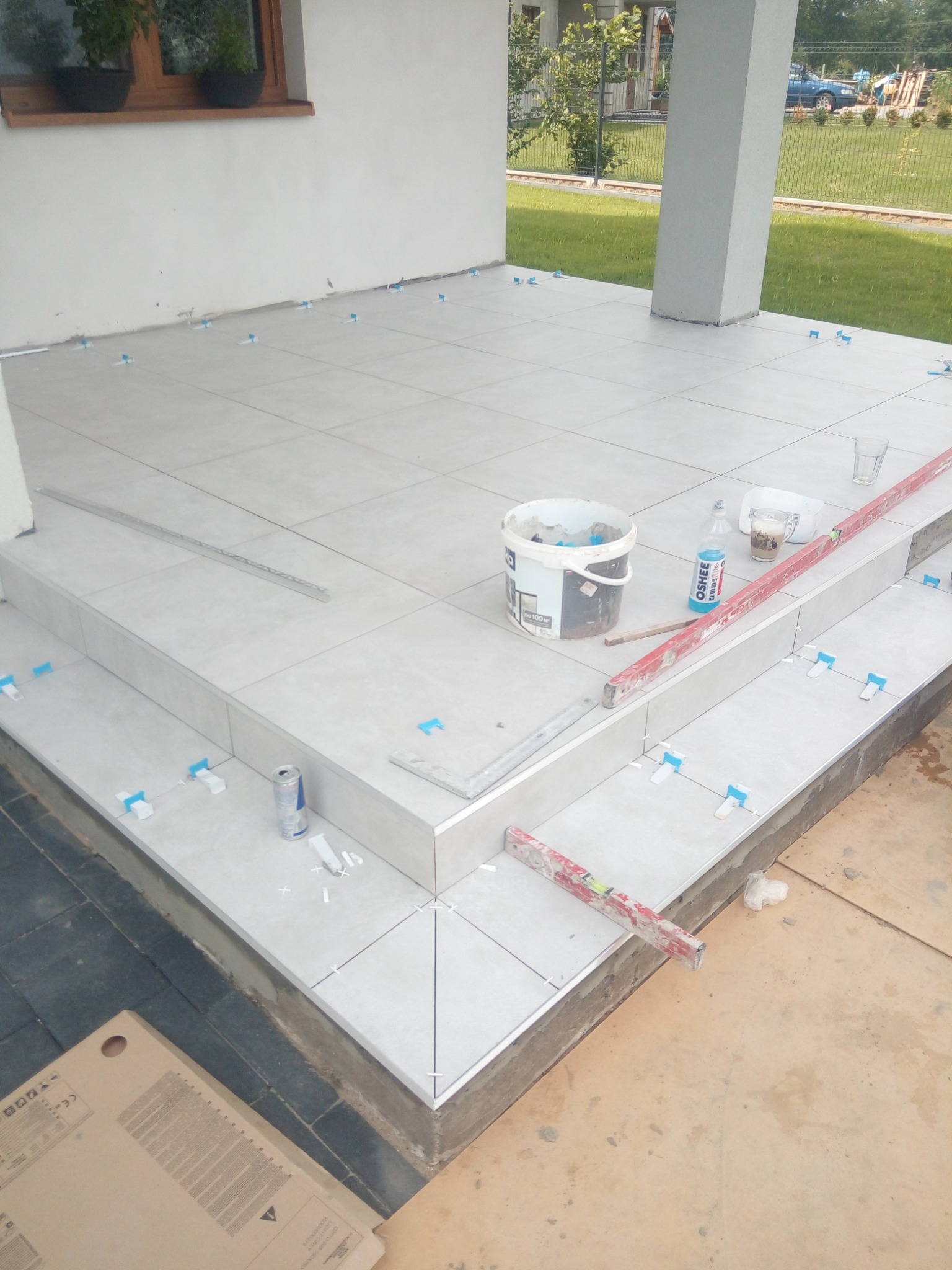 Układanie jasnych płytek na schodach wejściowych, widoczne poziomice, puszka z napojem, wiadro z zaprawą i elementy do poziomowania płytek.