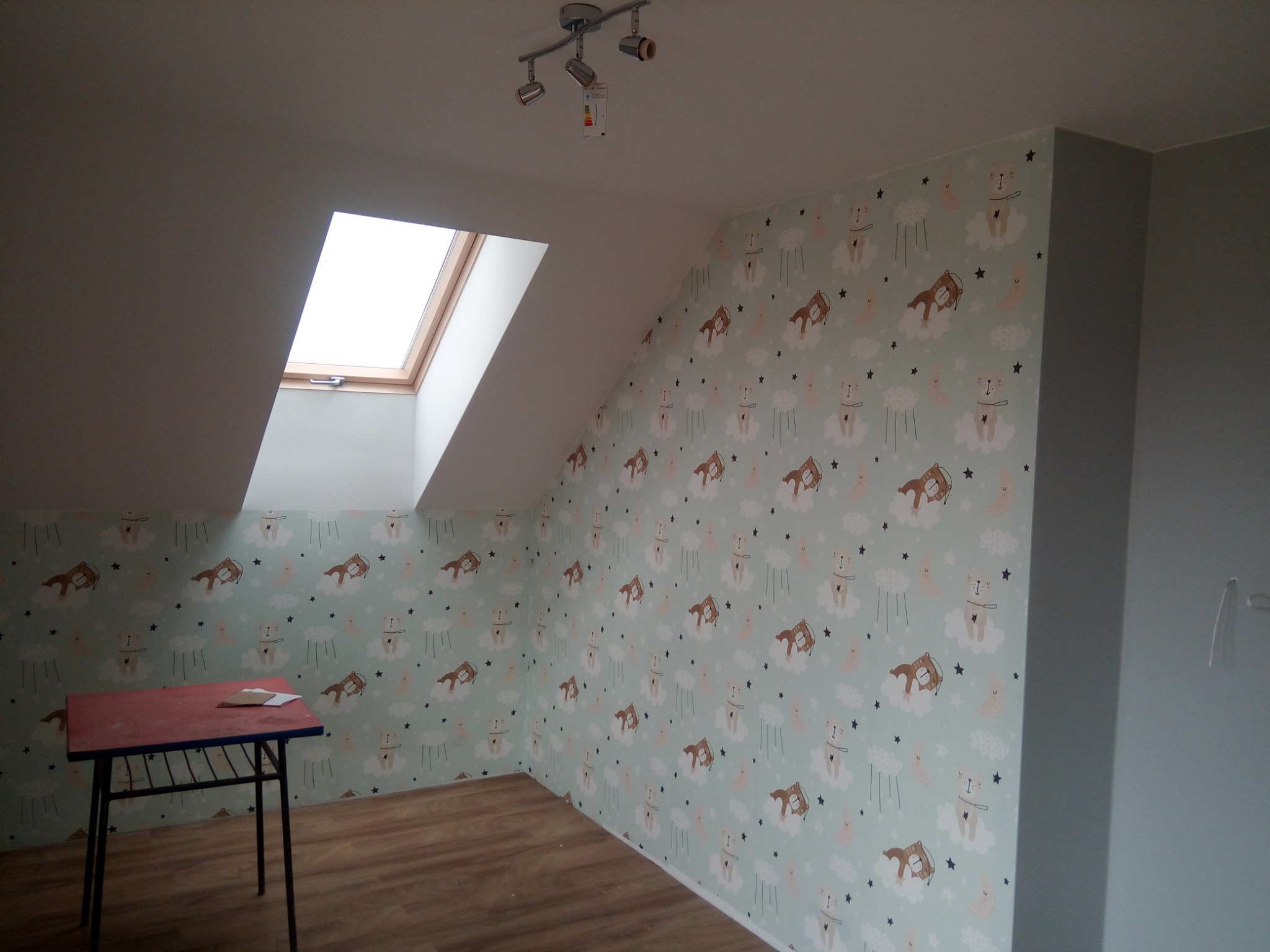 Pokój na poddaszu po odświeżeniu, ściany pokryte tapetą w dziecięce wzory (misie, konie na chmurach, gwiazdki), widoczne okno dachowe i lampa sufitowa.