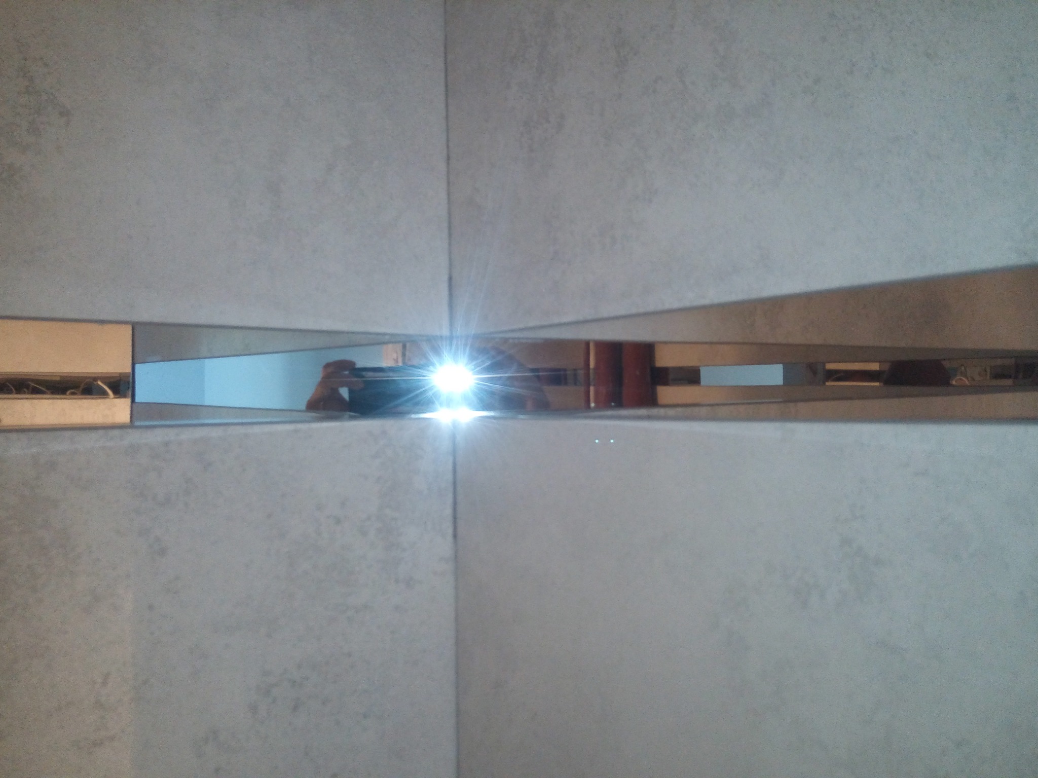 Fragment ściany wykończonej szarymi płytkami z lustrzanymi wstawkami, odbijającymi światło i sylwetkę osoby robiącej zdjęcie.