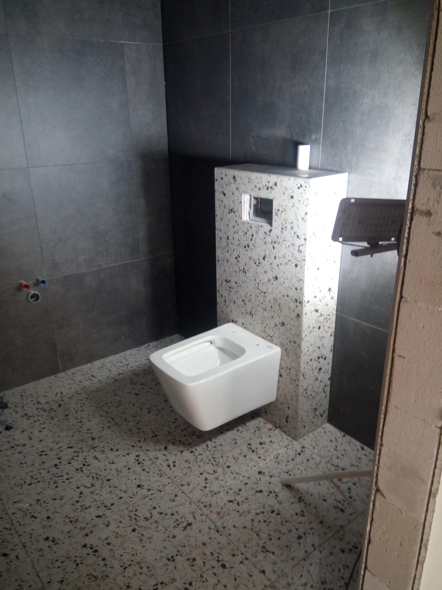 Montaż podwieszanej miski WC na stelażu w łazience z szarymi płytkami ściennymi i podłogą lastryko, widoczne przyłącza wodne i fragment surowej ściany.