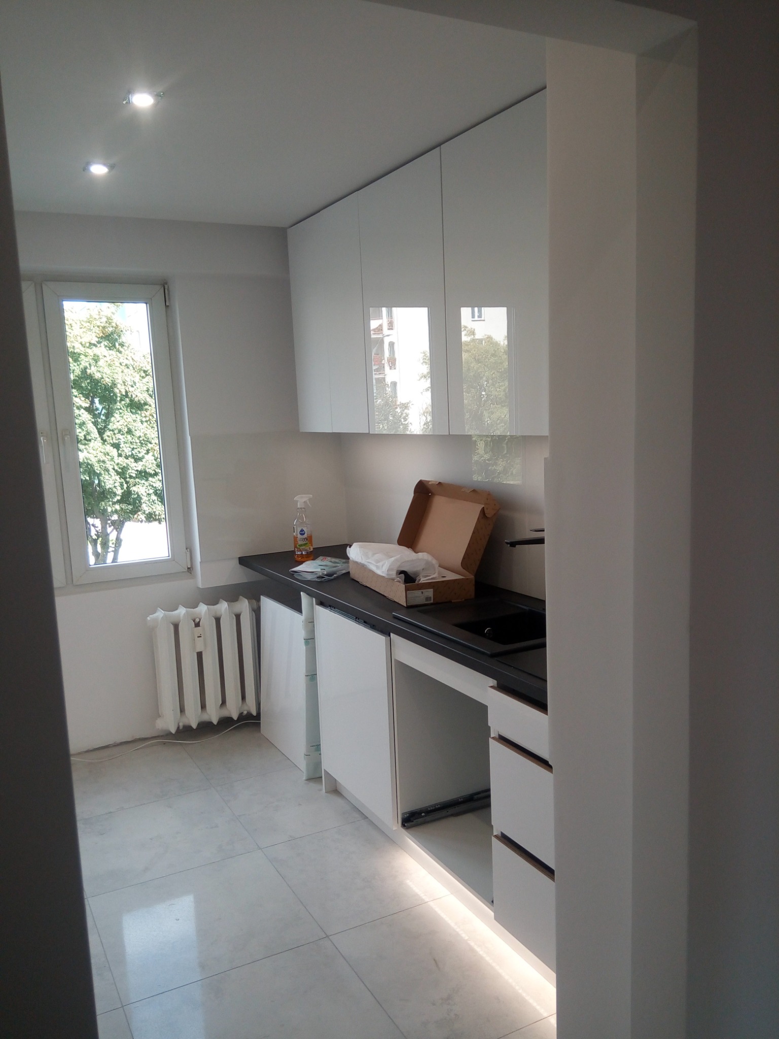 Nowoczesna kuchnia w trakcie montażu, białe szafki z połyskiem, czarny blat, widoczny karton z akcesoriami i środki czystości na blacie, płytki podłogowe, oświetlenie podszafkowe i grzejnik pod oknem.