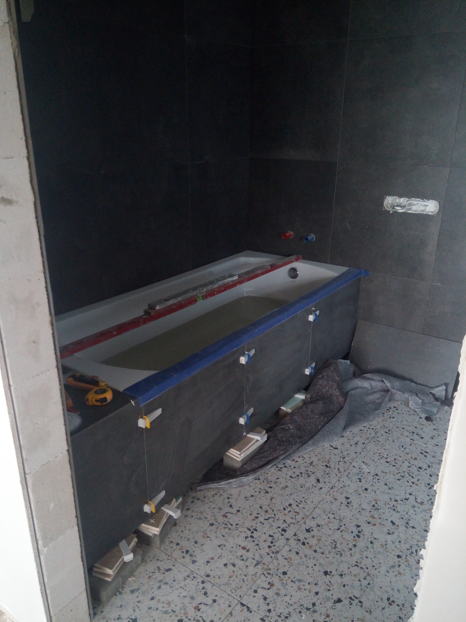 Montaż obudowy wanny z ciemnych płytek, poziomica i kliny stabilizujące, widoczna instalacja wodna na ścianie, podłoga z lastryko.