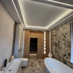 Nowoczesna łazienka z podświetleniem LED na suficie, marmurową ścianą i podłogą. Widoczna wanna, umywalka i toaleta. Oświetlenie w kształcie ramy dodaje elegancji.