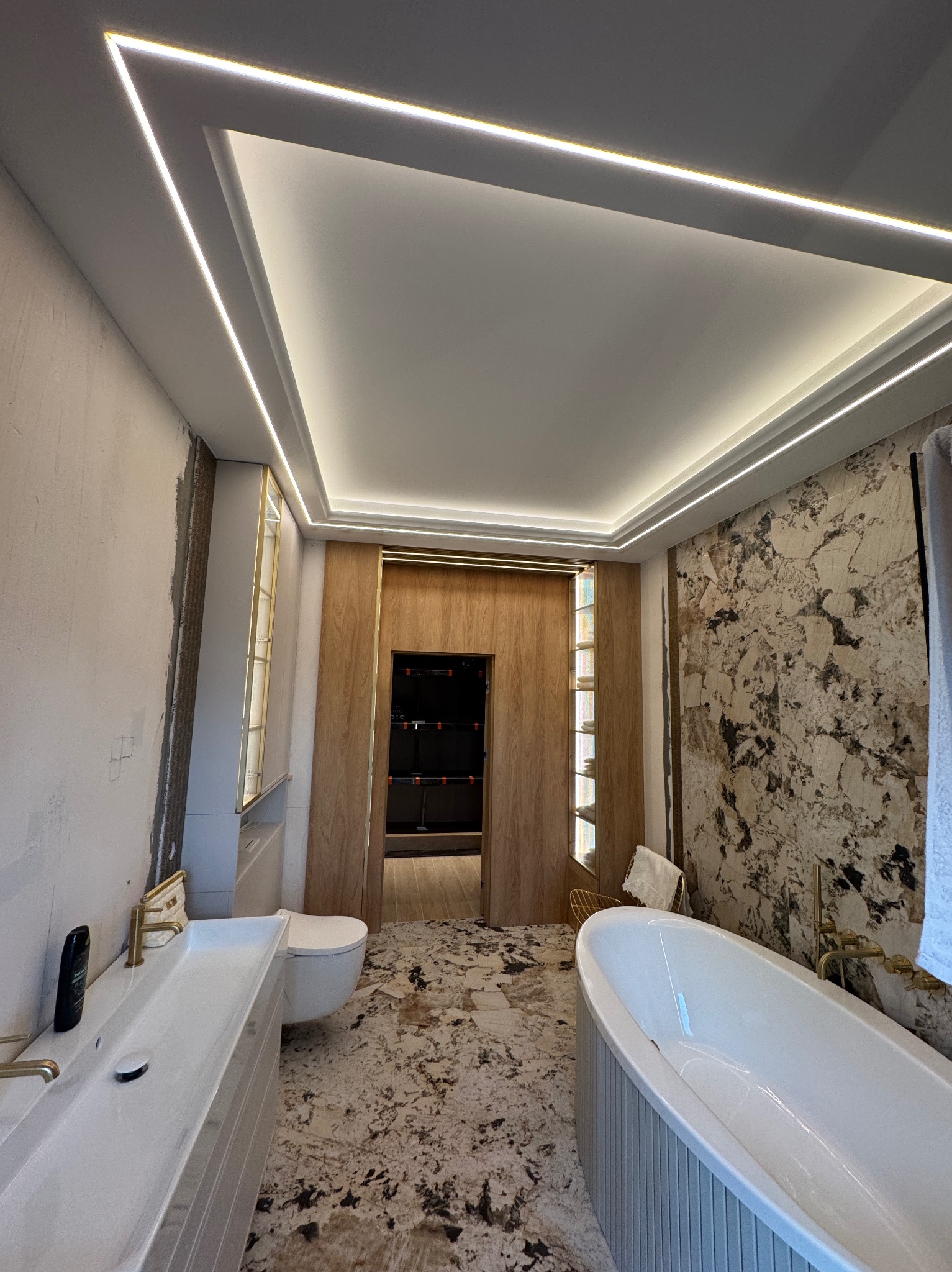 Nowoczesna łazienka z podświetleniem LED na suficie, marmurową ścianą i podłogą. Widoczna wanna, umywalka i toaleta. Oświetlenie w kształcie ramy dodaje elegancji.