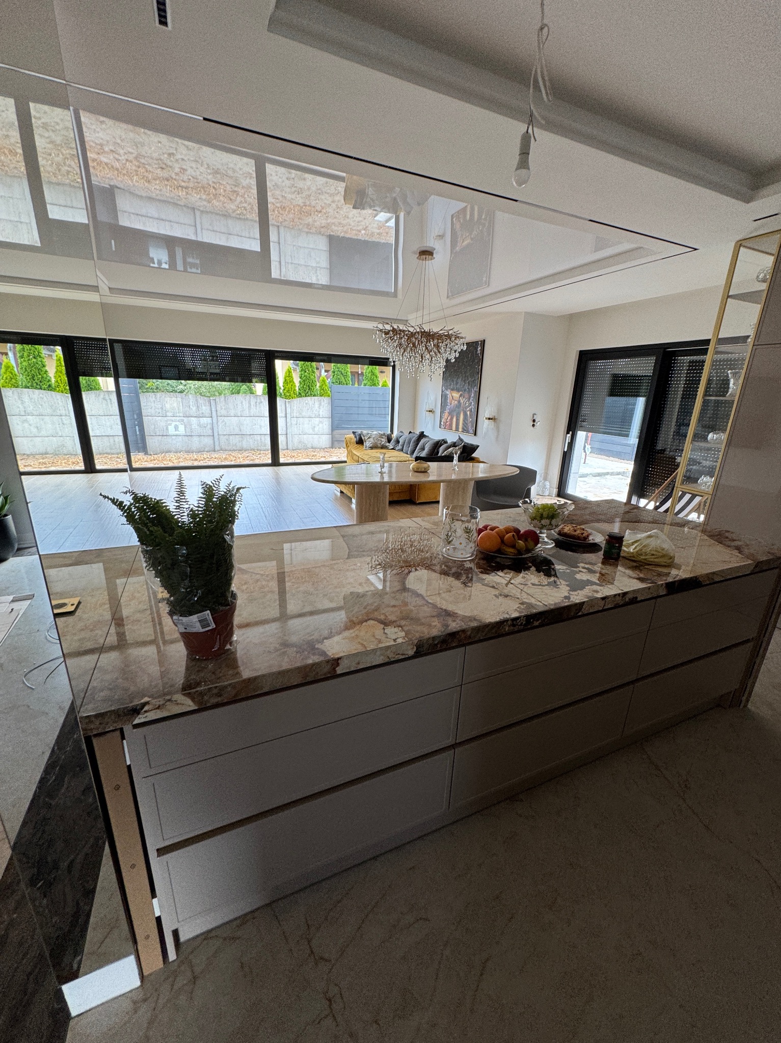 Nowoczesna kuchnia z wyspą i marmurowym blatem, widok na salon i okno.  W tle zwisający przewód z żarówką sugerujący montaż oświetlenia.