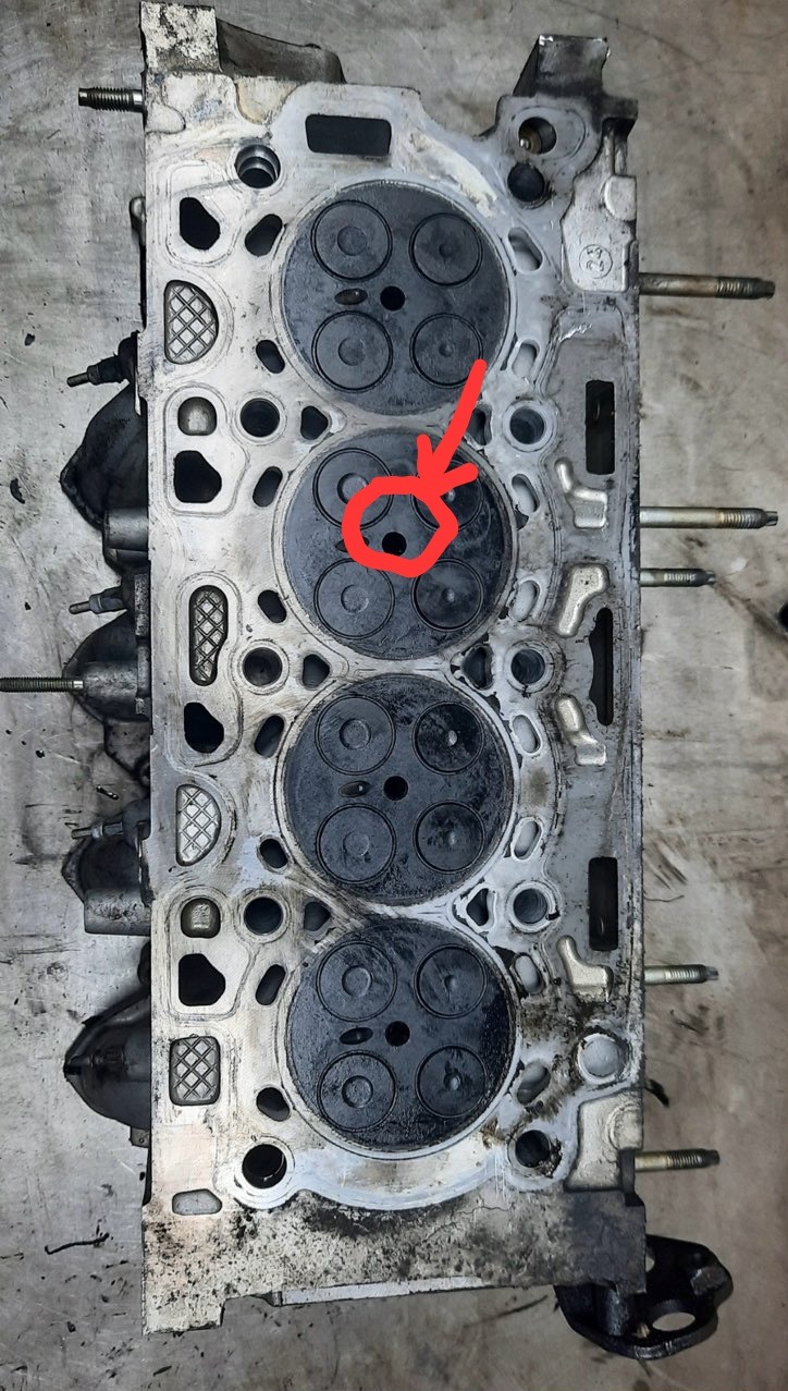 Głowica silnika samochodowego z zaznaczonym obszarem, prawdopodobnie po demontażu, widoczne ślady oleju i nagaru. Detal mechaniczny w warsztacie.