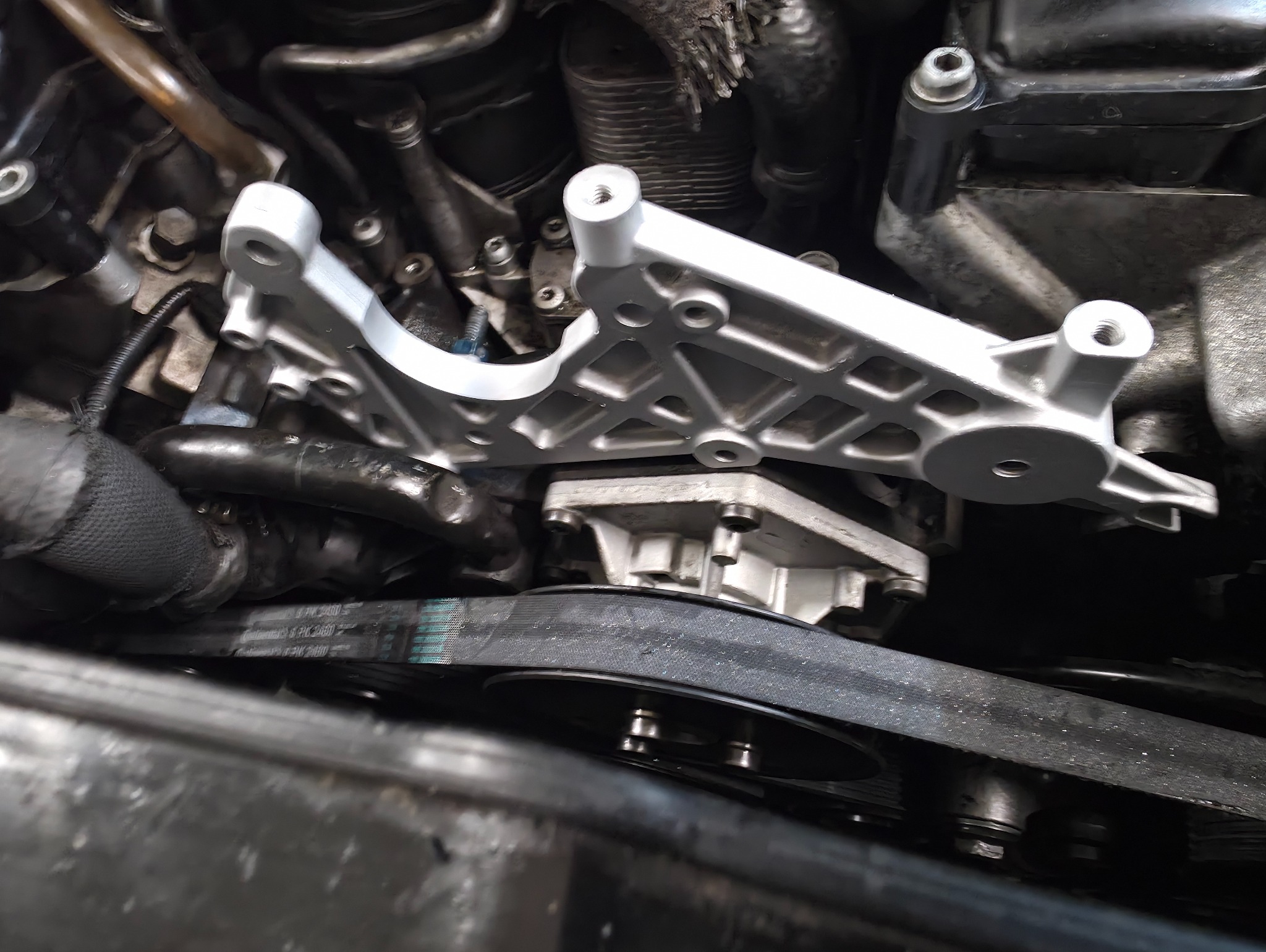 Szczegółowe ujęcie silnika samochodowego z nową, srebrną podporą. Widoczny pasek klinowy i elementy układu chłodzenia. Oświetlenie warsztatowe podkreśla detale.