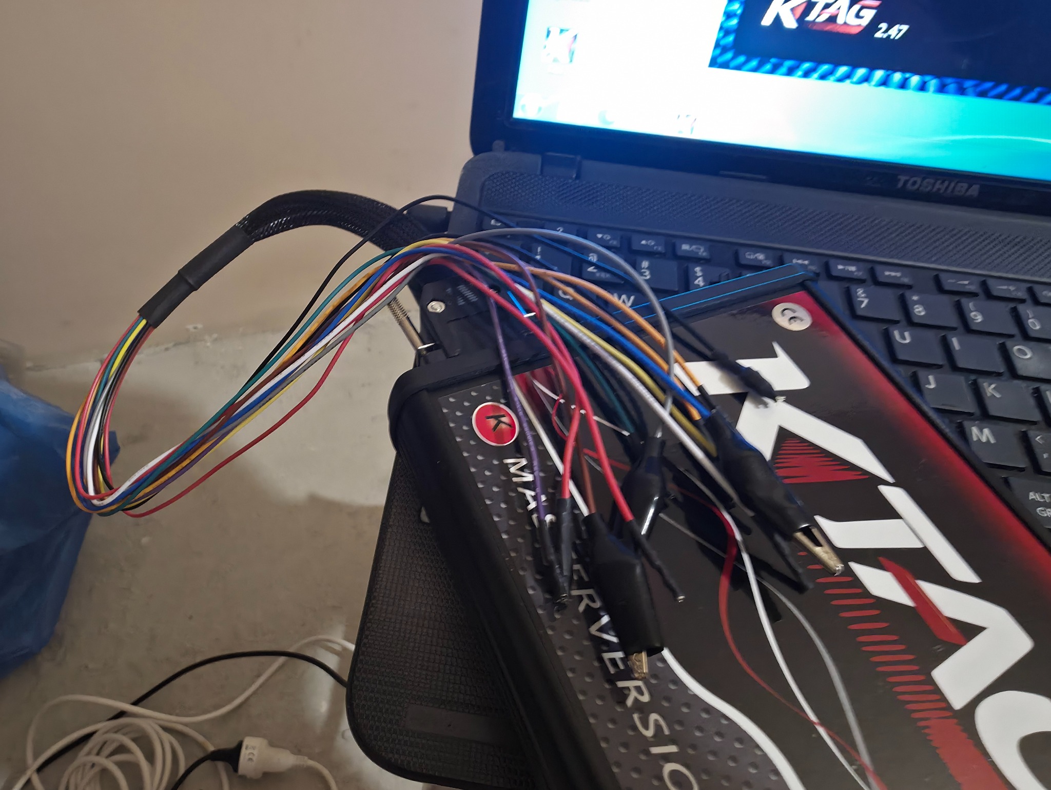 Interfejs diagnostyczny KTAG podłączony do laptopa, widoczne kolorowe przewody z krokodylkami. Sprzęt używany do tuningu samochodowego i diagnostyki komputerowej.