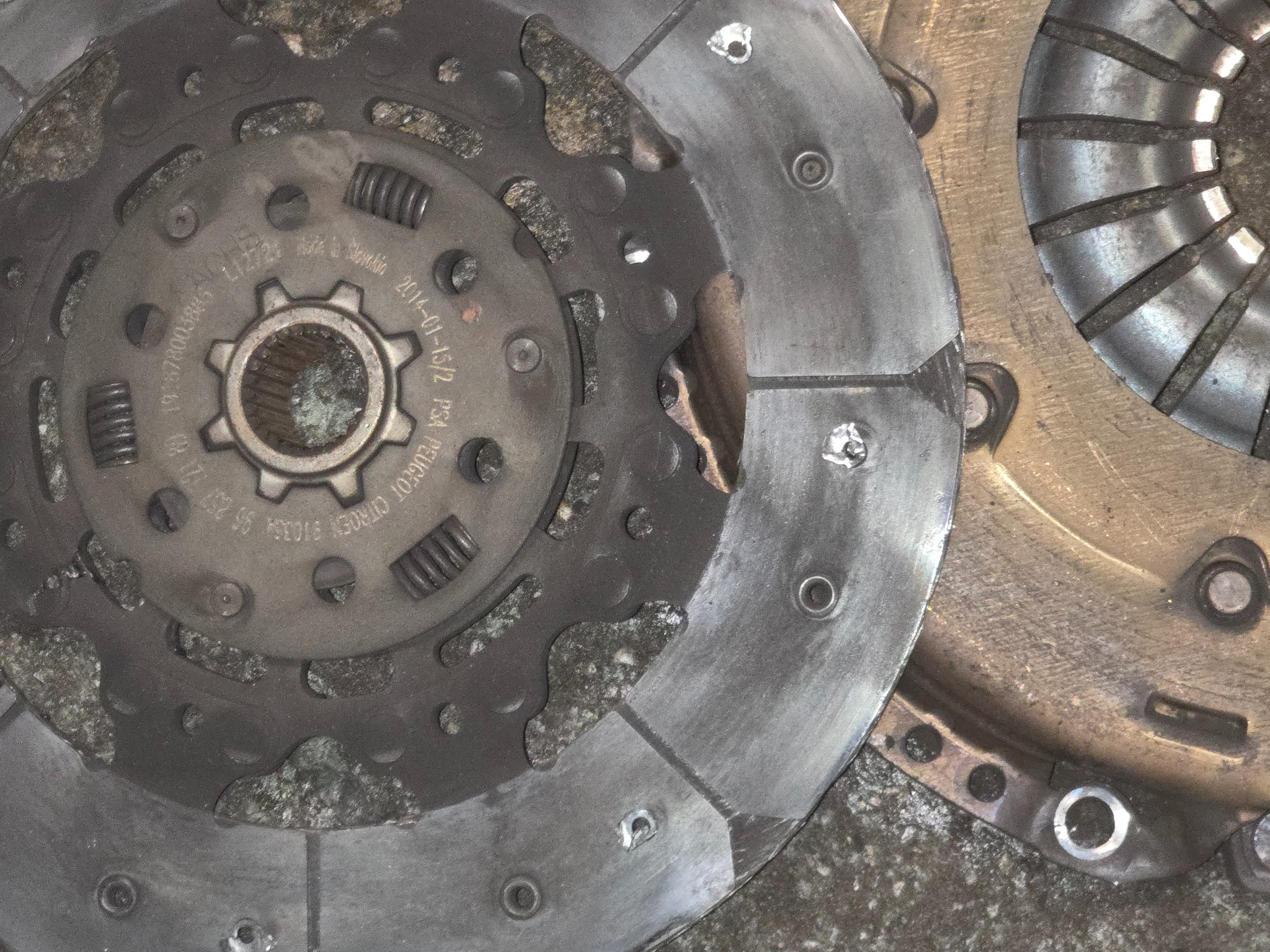 Zużyte tarcze sprzęgła Peugeot/Citroen z widocznymi śladami eksploatacji i numerami seryjnymi, leżące na szorstkiej, betonowej powierzchni. Detaliczny widok.