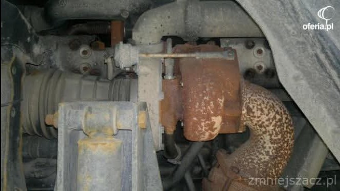 Zardzewiała turbosprężarka w samochodzie, widoczna od spodu, z elementami układu wydechowego i zawieszenia. Widoczne ślady korozji i zużycia.