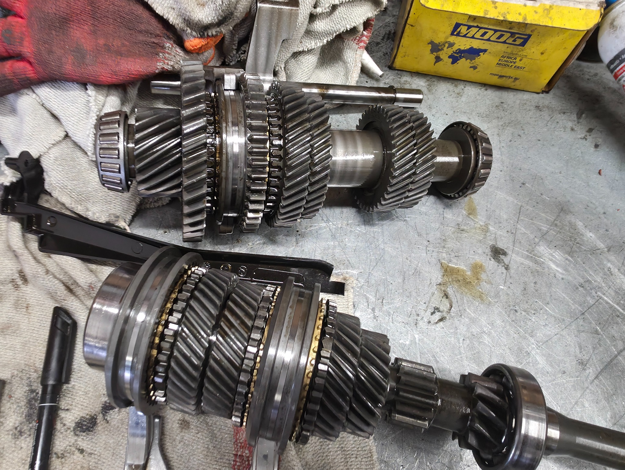 Dwa wały przekładni zębatych na metalowym stole warsztatowym, obok narzędzia i pudełko MOOG, ukazujące etap naprawy lub konserwacji skrzyni biegów.