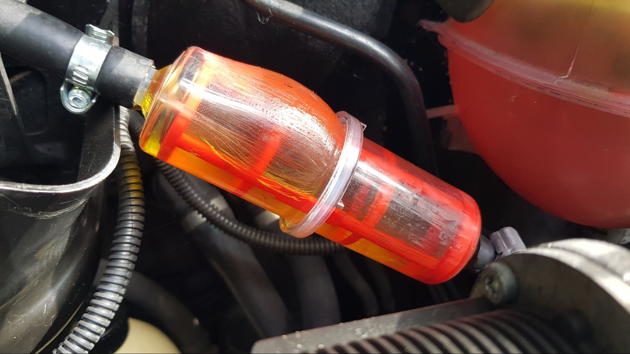 Przezroczysty filtr paliwa z pomarańczowo-czerwonym wkładem, zamontowany w przewodzie paliwowym silnika w Pomiechówku, zabezpieczony opaskami zaciskowymi.