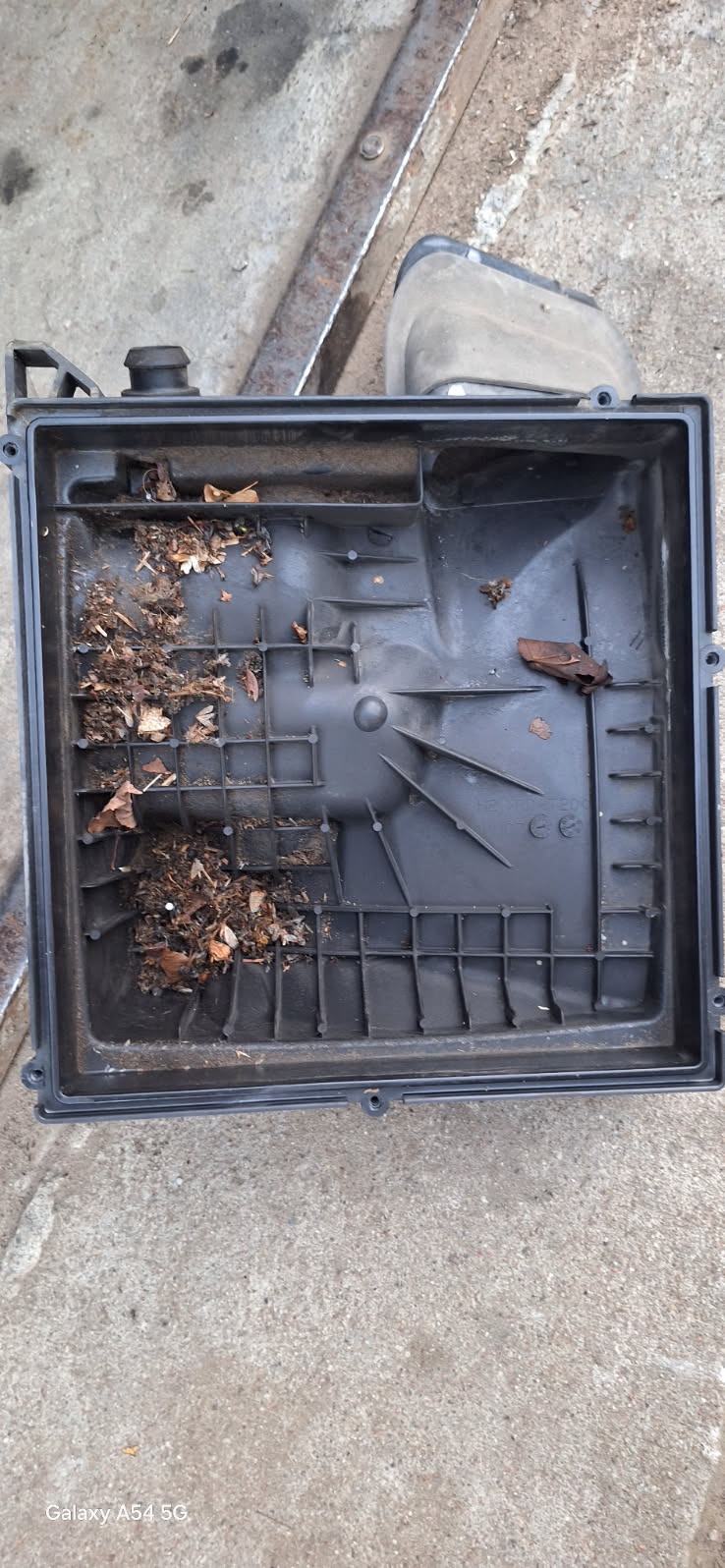 Otwarta, kwadratowa obudowa filtra powietrza z resztkami liści i brudu wewnątrz, leżąca na szarej, betonowej powierzchni. Widoczny fragment metalowej ramy w tle.