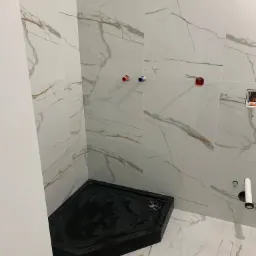 Czarny brodzik narożny w kabinie prysznicowej wyłożonej białymi płytkami imitującymi marmur z widocznymi przyłączami wodno-kanalizacyjnymi i elektrycznymi.