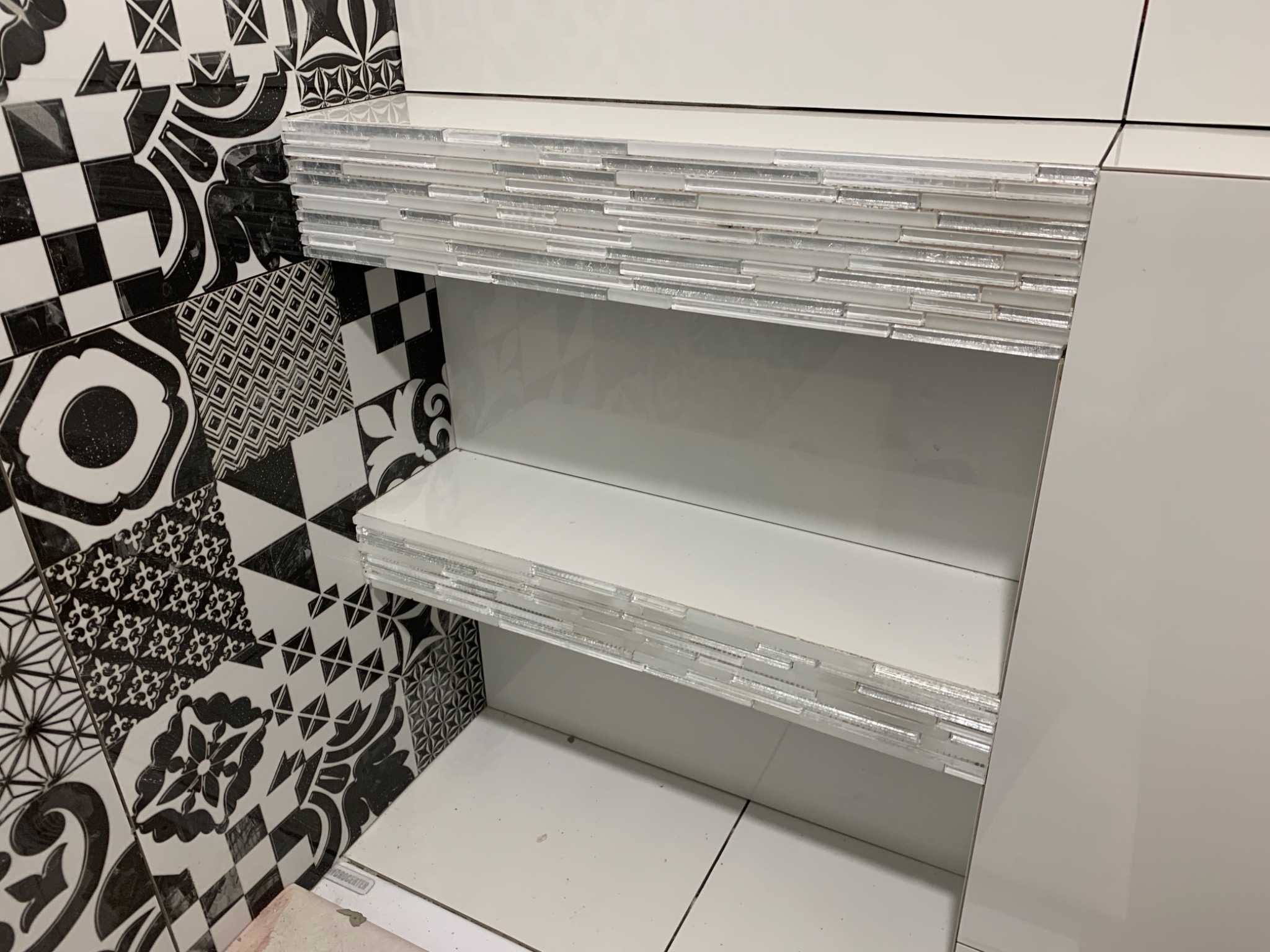 Wykończona wnęka w łazience z białymi półkami, obłożona mozaiką szklaną, z widoczną ścianą w czarno-białe, wzorzyste płytki.