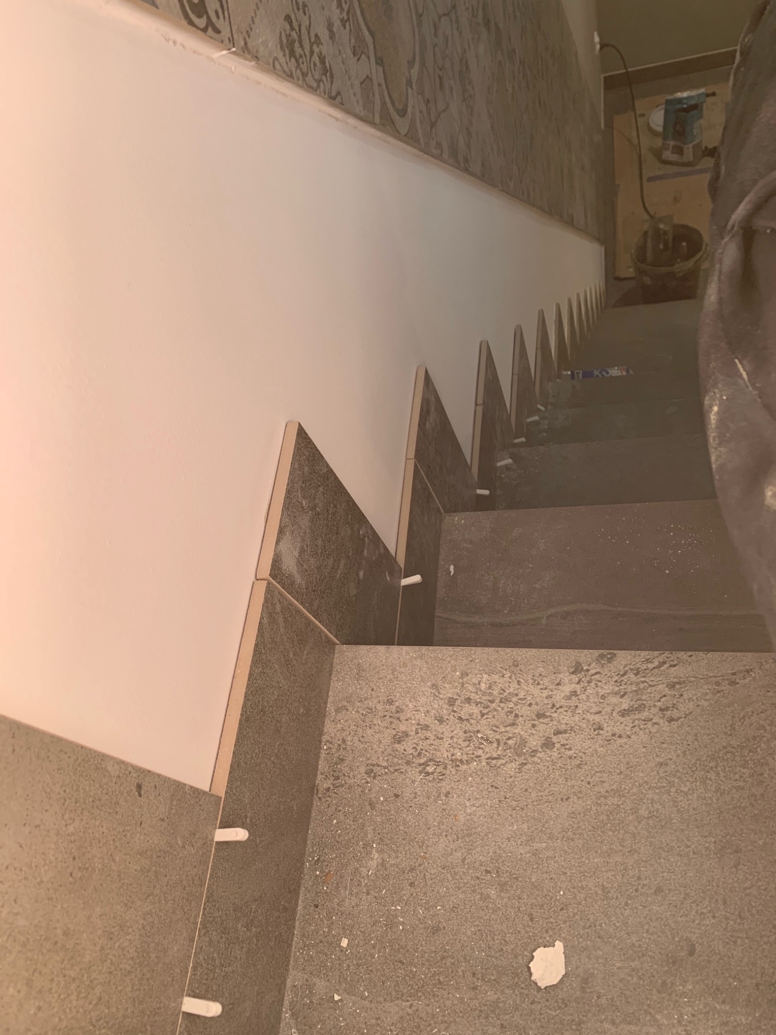 Montaż płytek na schodach w Łodzi, widoczne białe krzyżyki dystansowe, ściana pomalowana na biało, a na górze ściany wzorzysta tapeta.