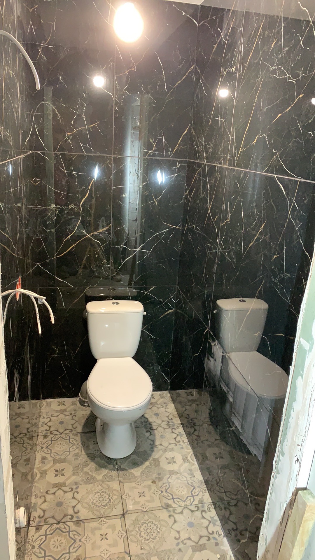 Wykończona łazienka z czarnymi płytkami imitującymi marmur i białym sedesem, podłoga wyłożona płytkami w stylu vintage, widoczne rurki instalacji wodnej.