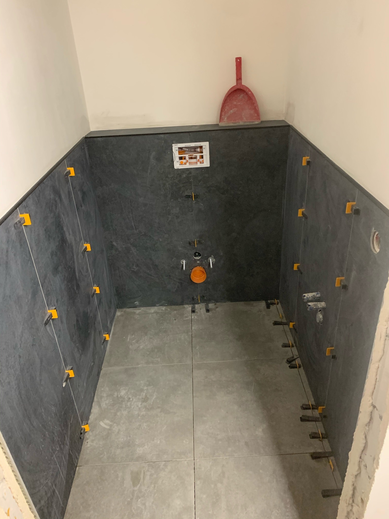 Montaż ciemnych płytek imitujących kamień na ścianach i jasnych płytek podłogowych w małej łazience, widoczne pomarańczowe poziomice krzyżowe, czerwona szufelka na górnej krawędzi ściany.