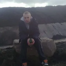 Mężczyzna siedzi na kamieniu z butelką w ręku na tle hałdy ziemi i pochmurnego nieba.