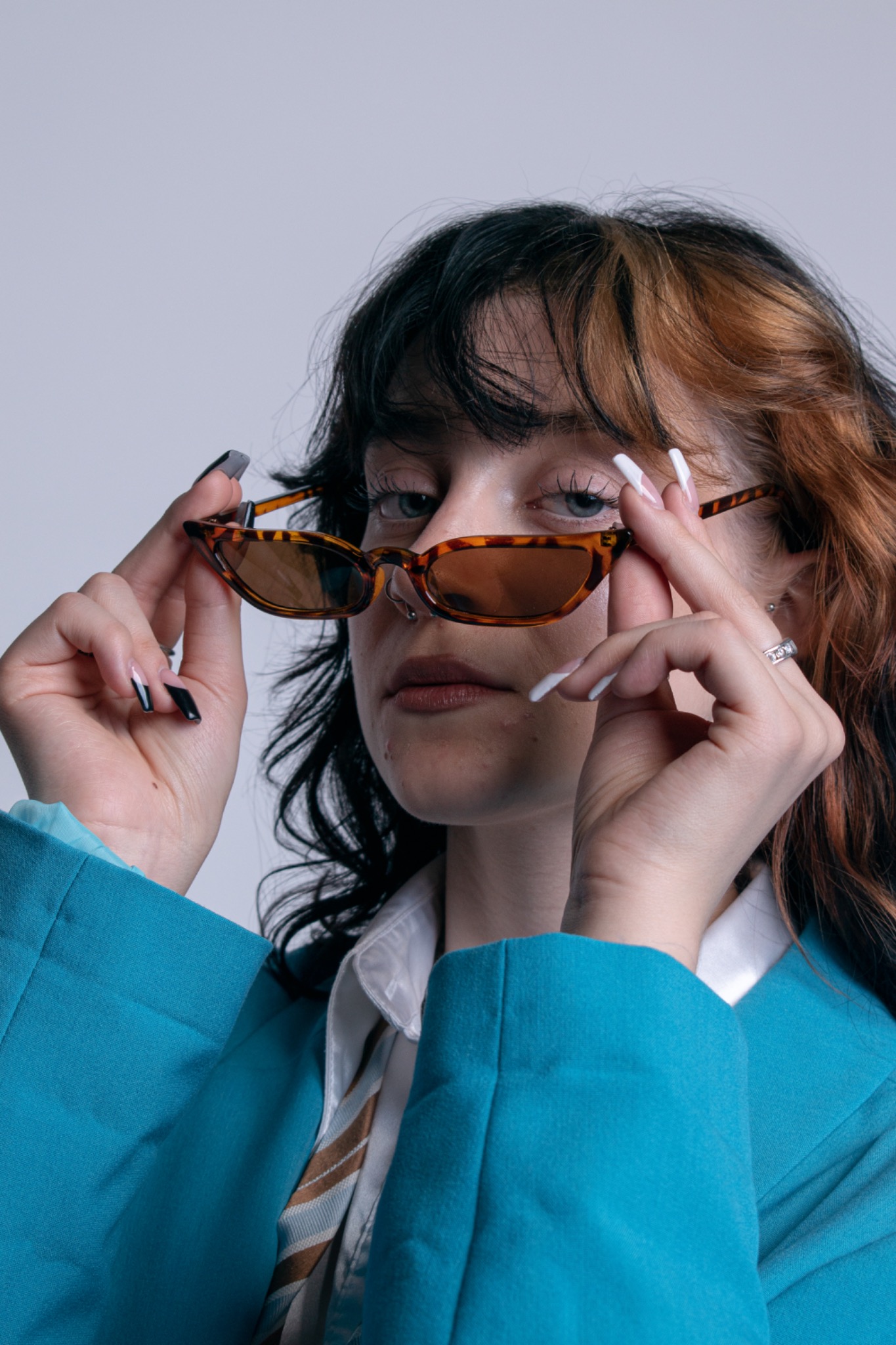Portret kobiety w turkusowej marynarce, trzymającej w dłoniach okulary w brązowej oprawce, z widocznymi piercingami w nosie, wykonany w Krakowie.