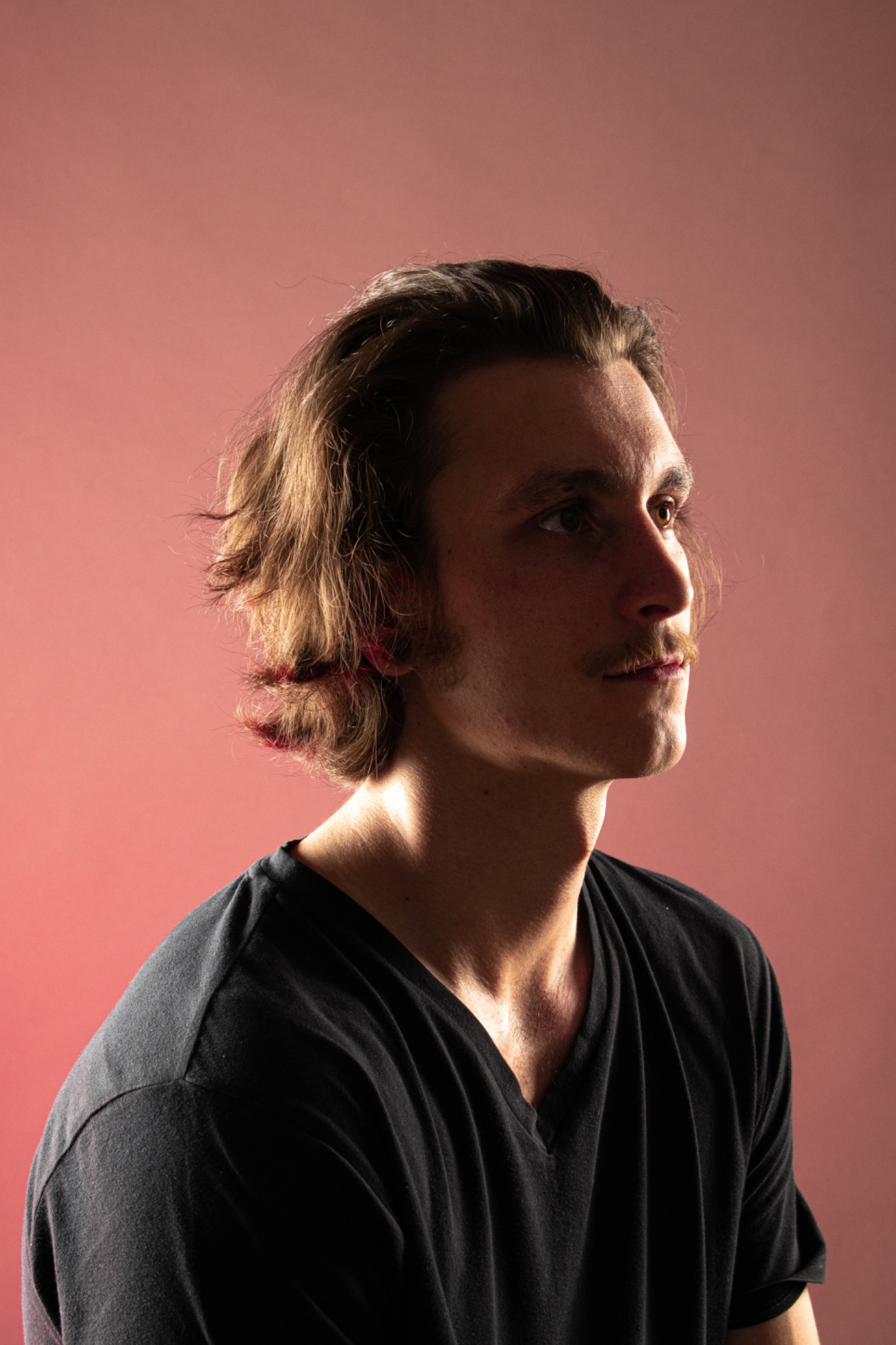 Portret młodego mężczyzny z wąsami, profil, brązowe włosy, czarna koszulka na tle różowego studia.
