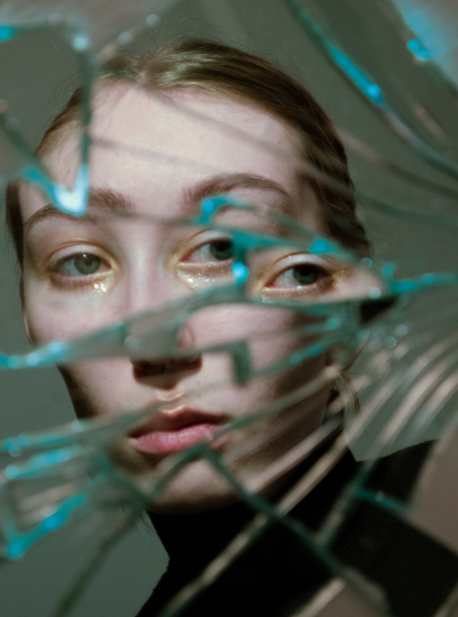 Portret kobiety z brokatem pod oczami, widzianej przez potłuczone szkło z niebieskimi refleksami.