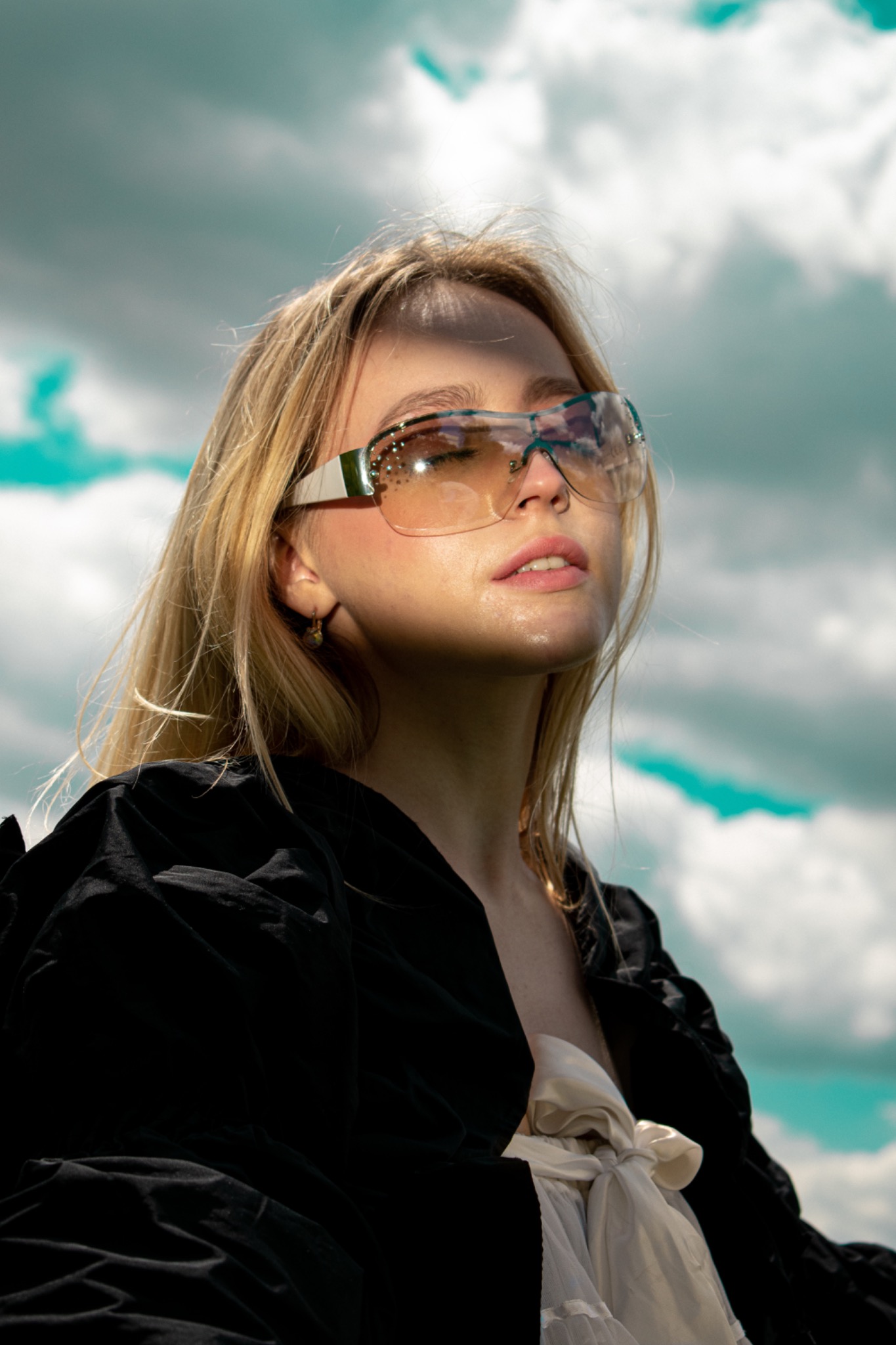 Portret kobiety z blond włosami, w okularach przeciwsłonecznych z ozdobnymi kamieniami, ubrana w czarną bluzkę i białą sukienkę, na tle błękitnego nieba z chmurami.