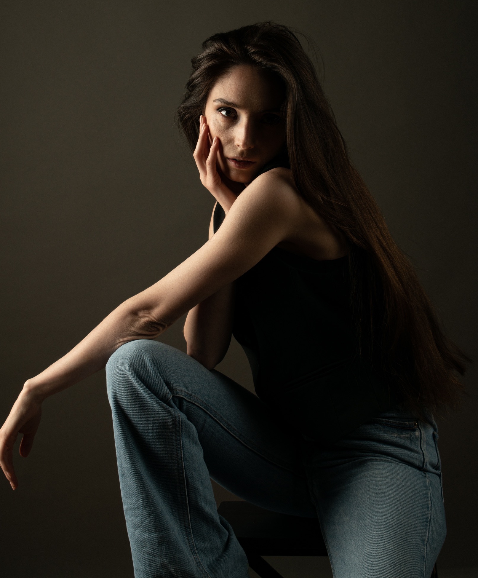 Portret kobiety z długimi, ciemnymi włosami, w czarnym topie i jeansach, siedzącej na krześle w ciemnym pomieszczeniu, z ręką opartą na twarzy, spoglądającej w obiektyw.
