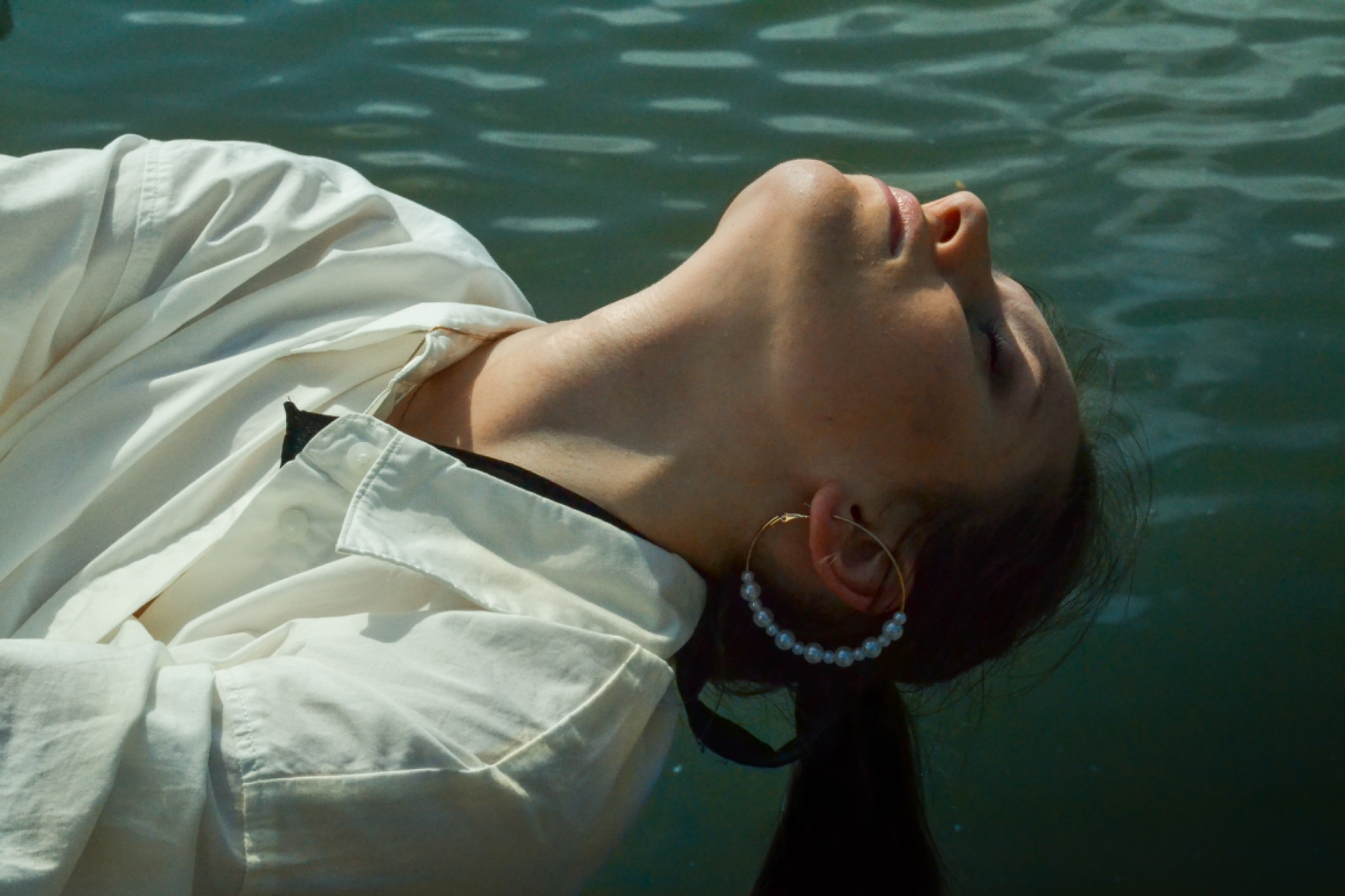 Kobieta w białej koszuli leżąca na wodzie, z głową odchyloną do tyłu, z kolczykiem kołem z perłami w uchu.