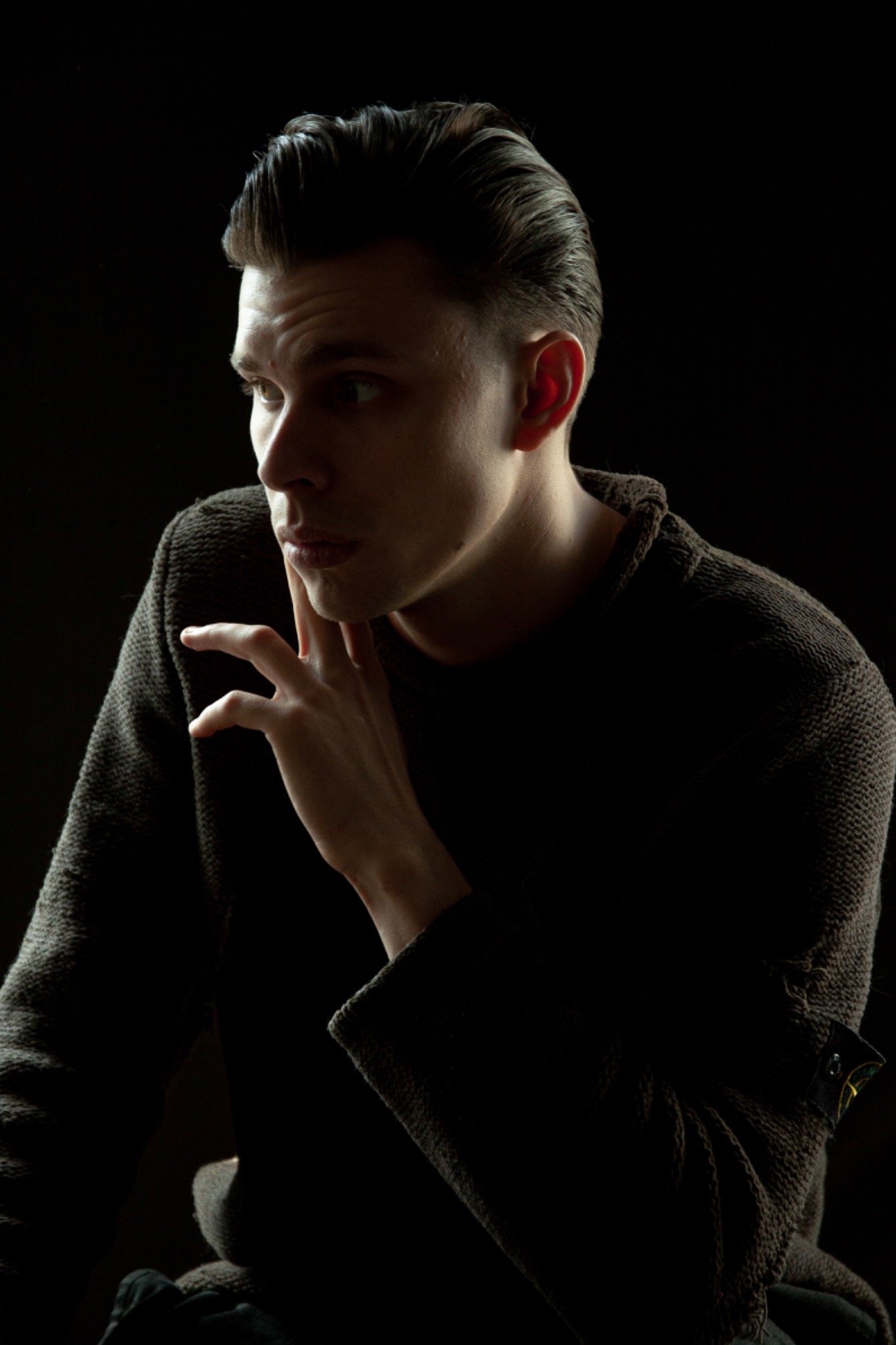 Portret mężczyzny w ciemnym swetrze, z ręką przy brodzie, na czarnym tle.