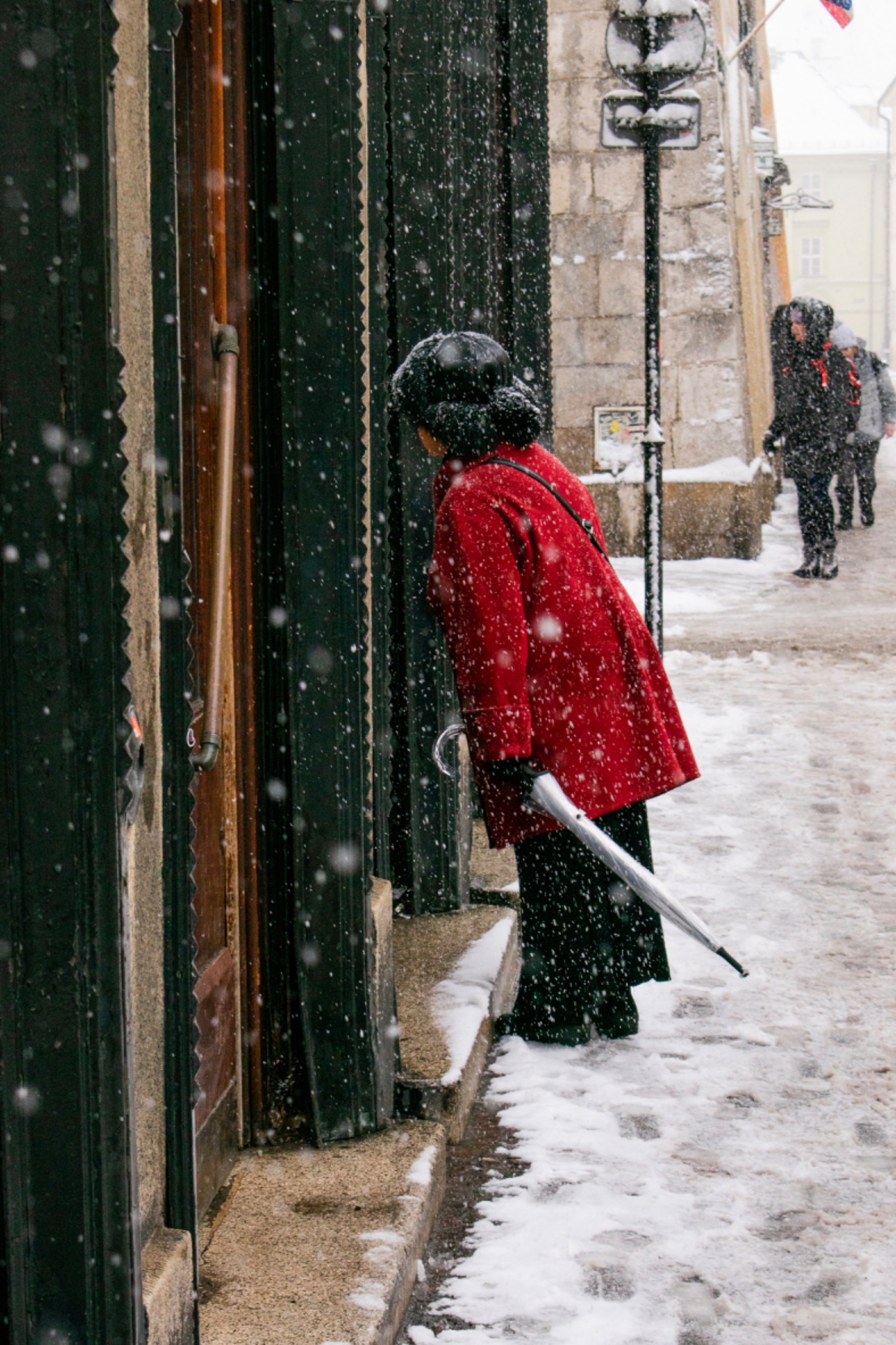 Kobieta w czerwonym płaszczu z parasolem, spogląda przez okno w śnieżny dzień. W tle przechodnie.