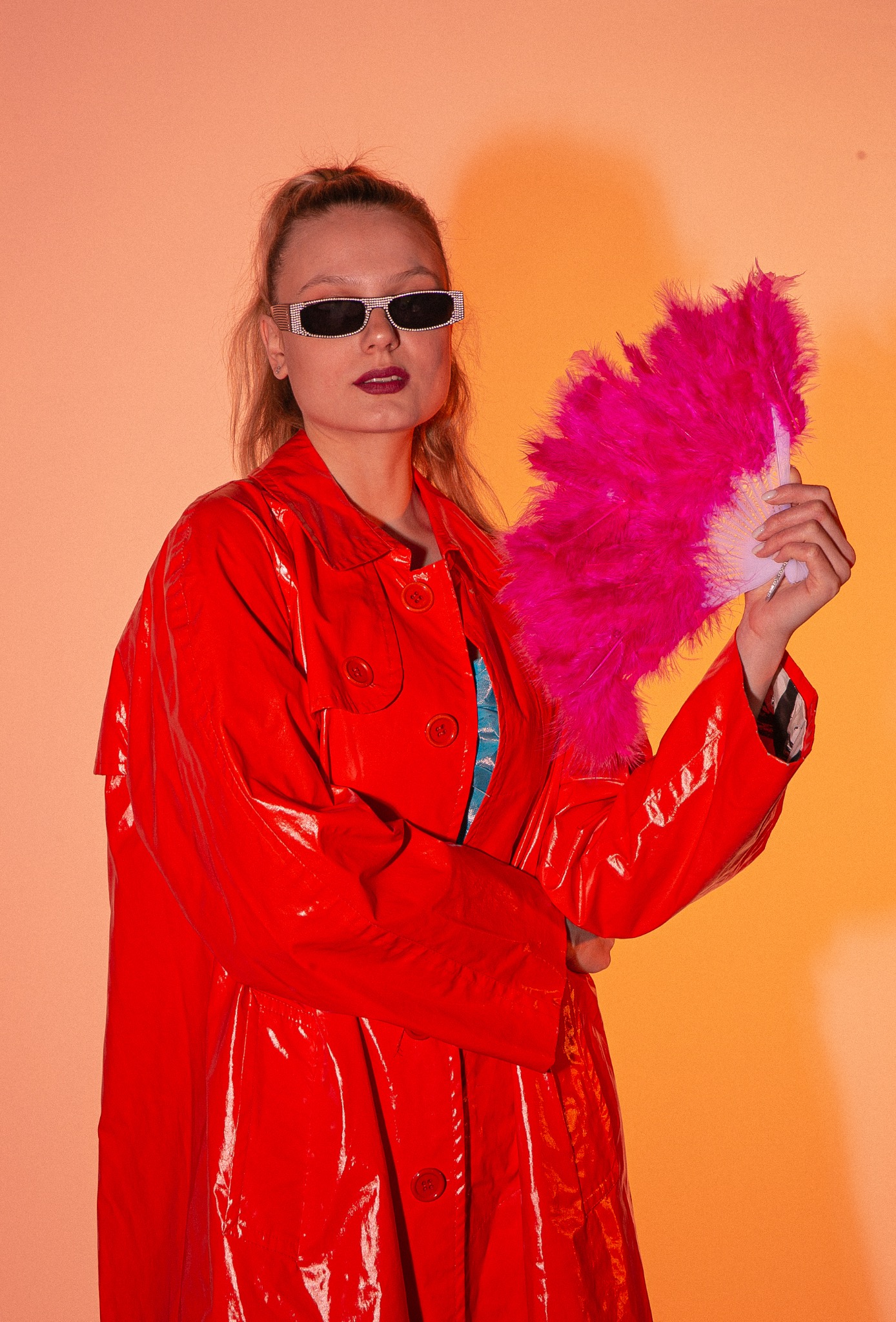 Kobieta w czerwonym, błyszczącym płaszczu i okularach z cyrkoniami, trzymająca różowy wachlarz z piór na pomarańczowym tle.