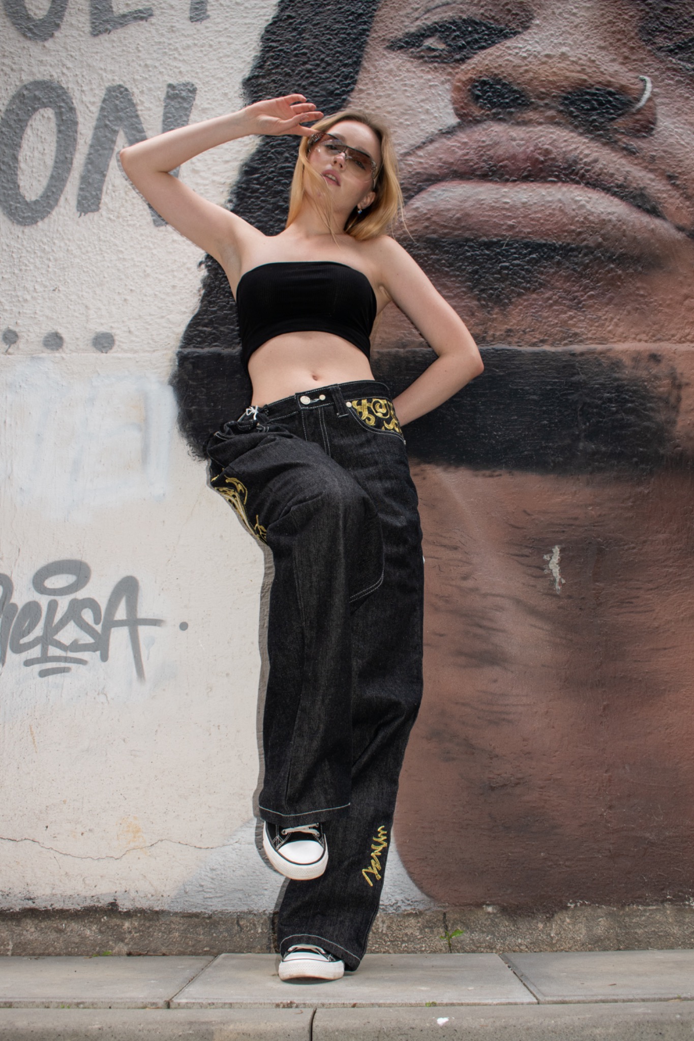 Młoda kobieta w czarnym topie i szerokich jeansach z haftem, pozująca na tle ściany z graffiti i częściowo widocznym portretem twarzy.