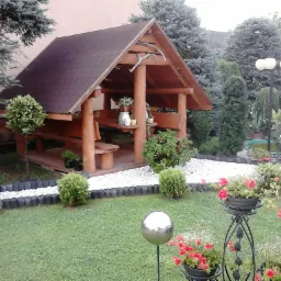 Drewniana altana ogrodowa z brązowym dachem, ozdobiona porożem, otoczona zadbanym trawnikiem, rabatami z kwiatami i dekoracyjnymi kulami