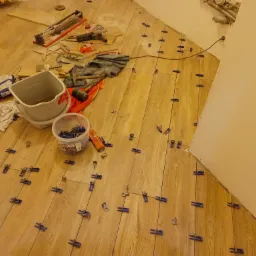 Układanie płytek podłogowych imitujących drewno z użyciem systemu poziomowania płytek; widoczne narzędzia glazurnicze, wiadro i klej.