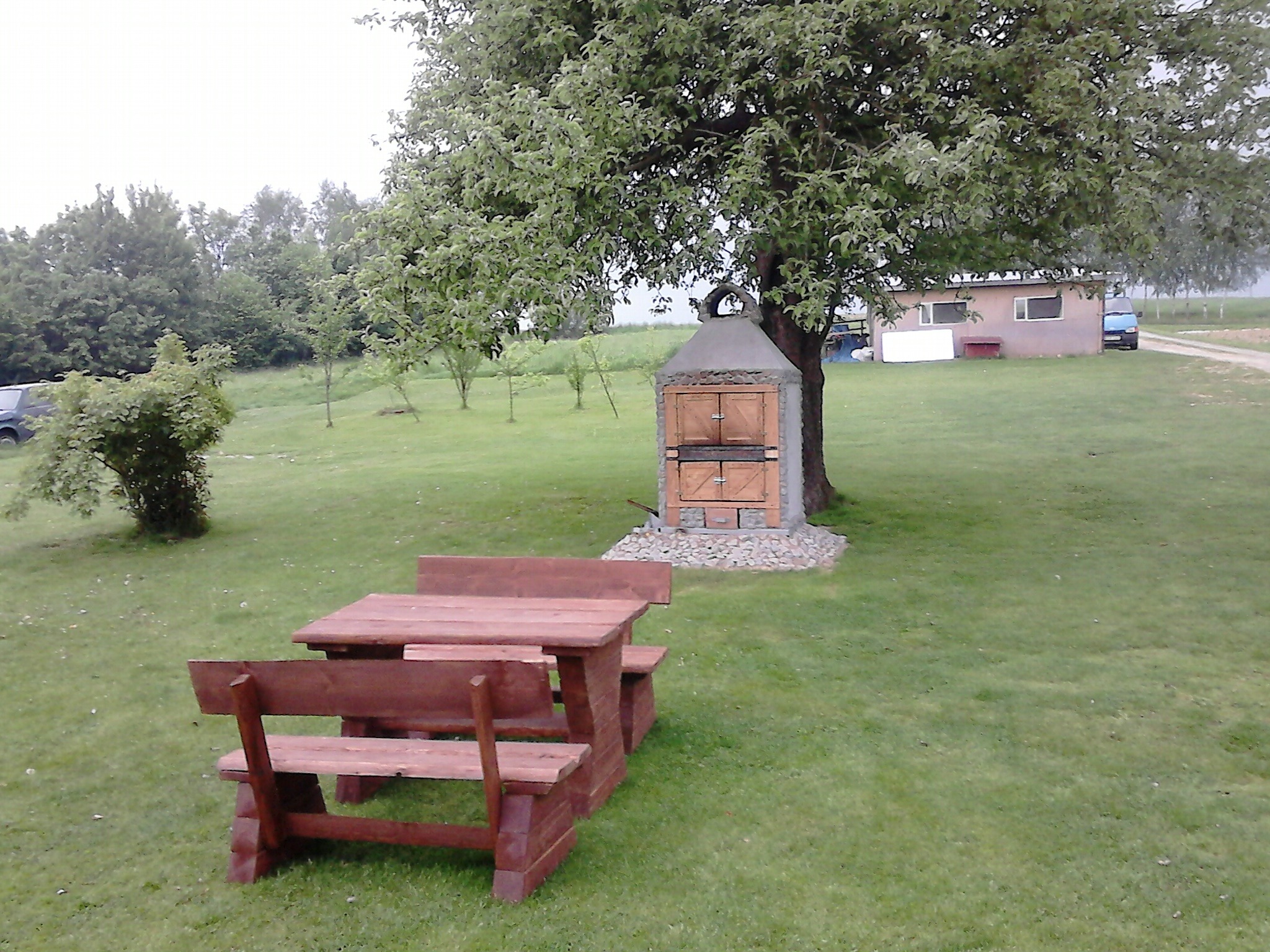 Drewniany stół piknikowy z ławkami na trawie, w tle murowany grill pod drzewem i budynek gospodarczy.