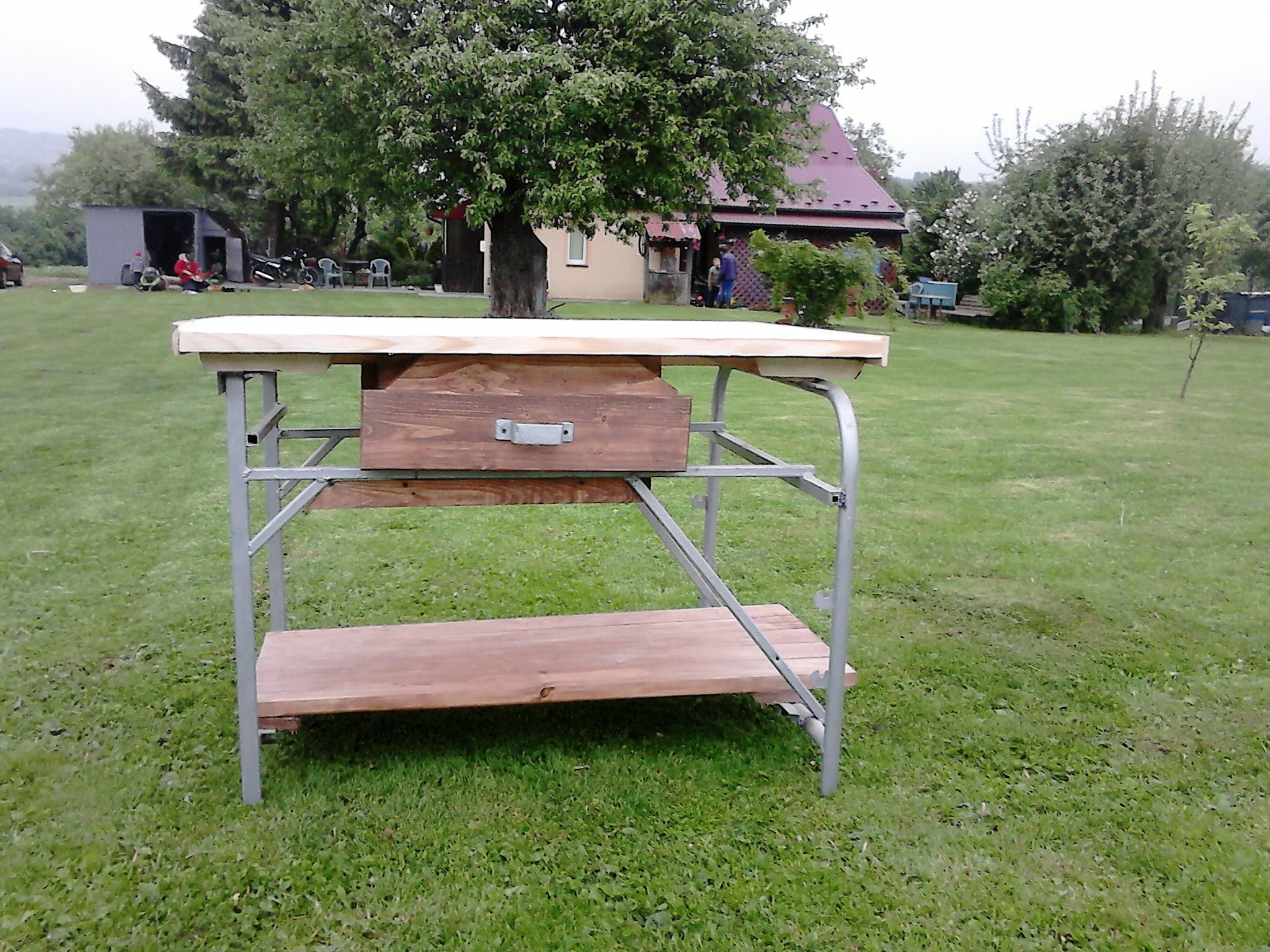 Stół roboczy z drewnianym blatem i metalową konstrukcją, z szufladą i półką, ustawiony na trawniku przed domem.