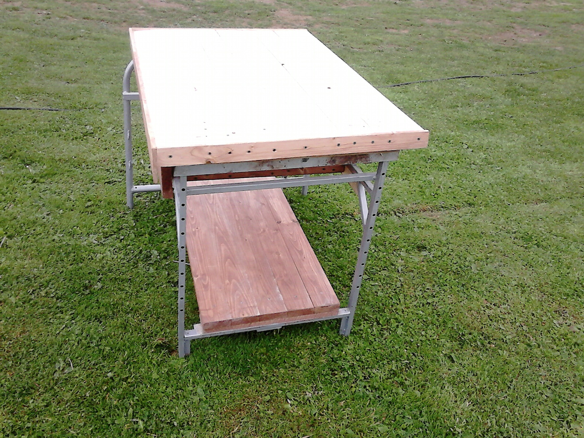 Stół warsztatowy z drewnianym blatem i metalową konstrukcją, stojący na trawie, widoczne ślady po wkrętach w blacie.