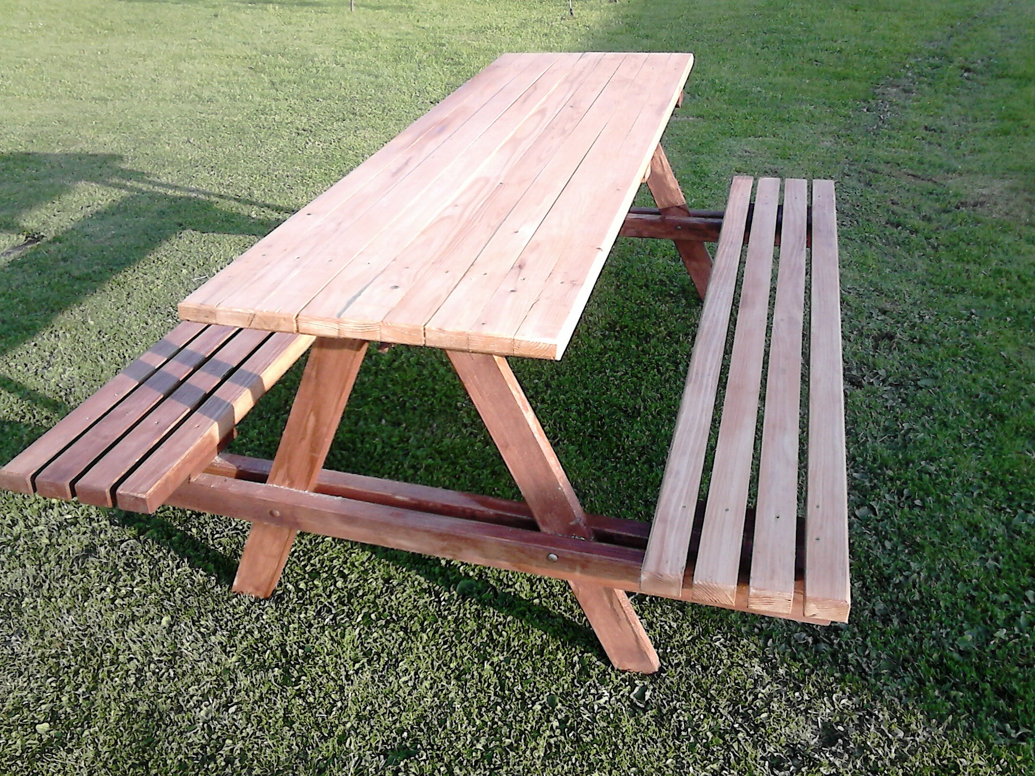 Drewniany stół piknikowy z ławkami na trawie, jedna ławka z ciemniejszego drewna, widoczne ślady po sękach.