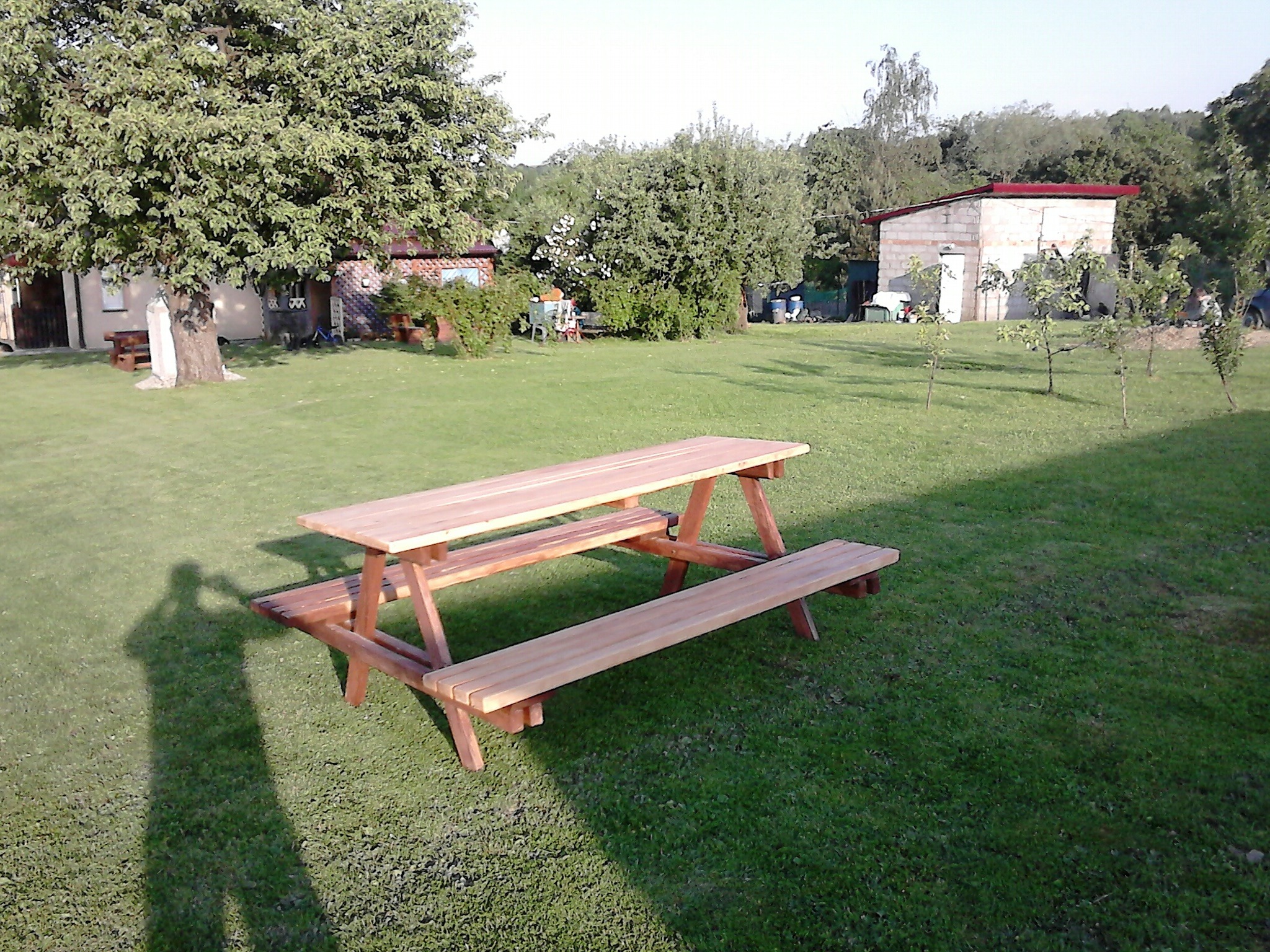 Nowy, drewniany stół piknikowy z ławkami na trawniku, w tle budynek gospodarczy i drzewa, widoczny cień fotografującego.