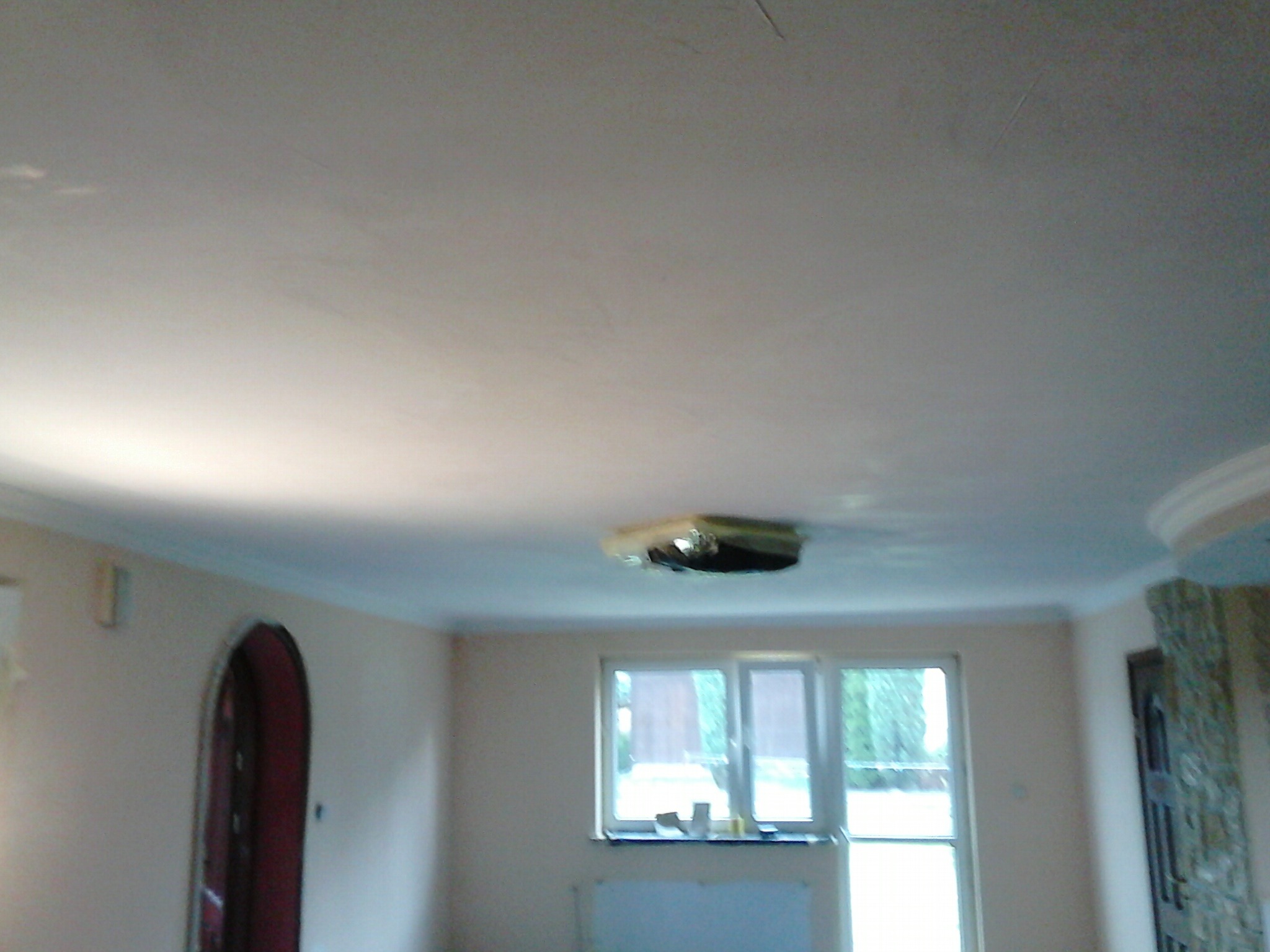 Pokój po remoncie: gładka, jasna ściana i sufit z widoczną instalacją oświetleniową, okno z widokiem na zieleń, dekoracyjny łuk drzwiowy i fragment ściany z elementami kamiennymi