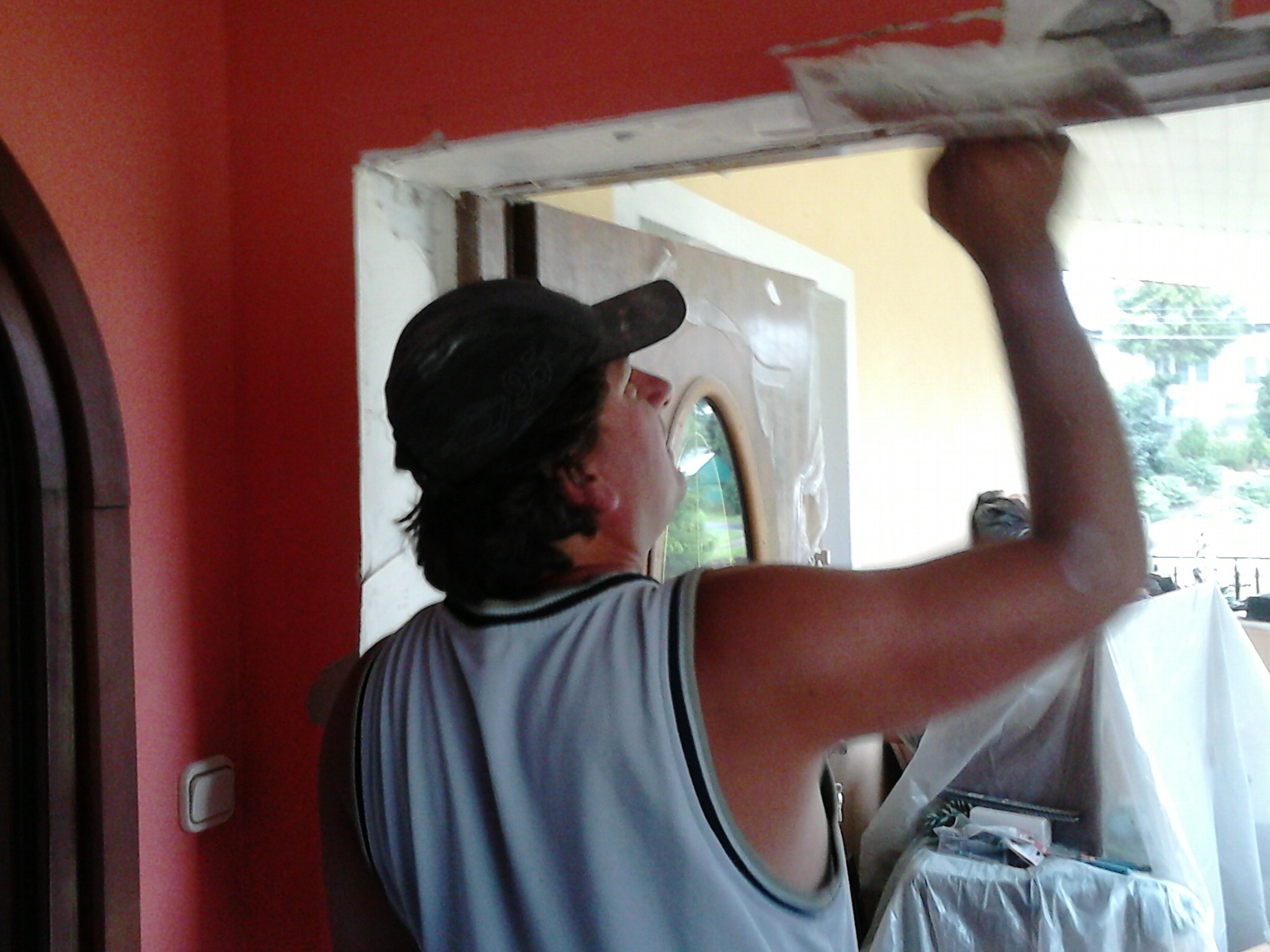 Mężczyzna w czapce wyrównuje szpachelką świeżo nałożoną masę szpachlową na ościeżnicy drzwi wewnętrznych, widoczna czerwona ściana i zabezpieczone folią meble.