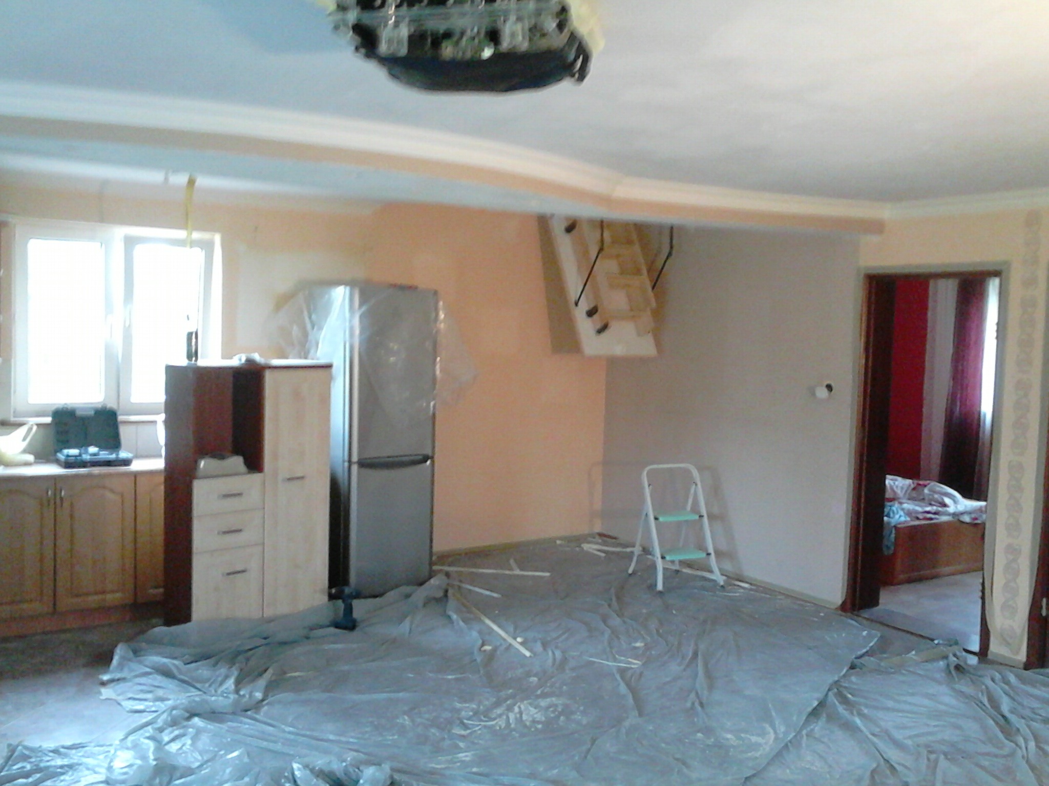 Pokój w trakcie malowania: podłoga zabezpieczona folią, widoczna drabina malarska, meble odsunięte i zabezpieczone, ściana częściowo pomalowana na jasny odcień, widoczne narzędzia malarskie.