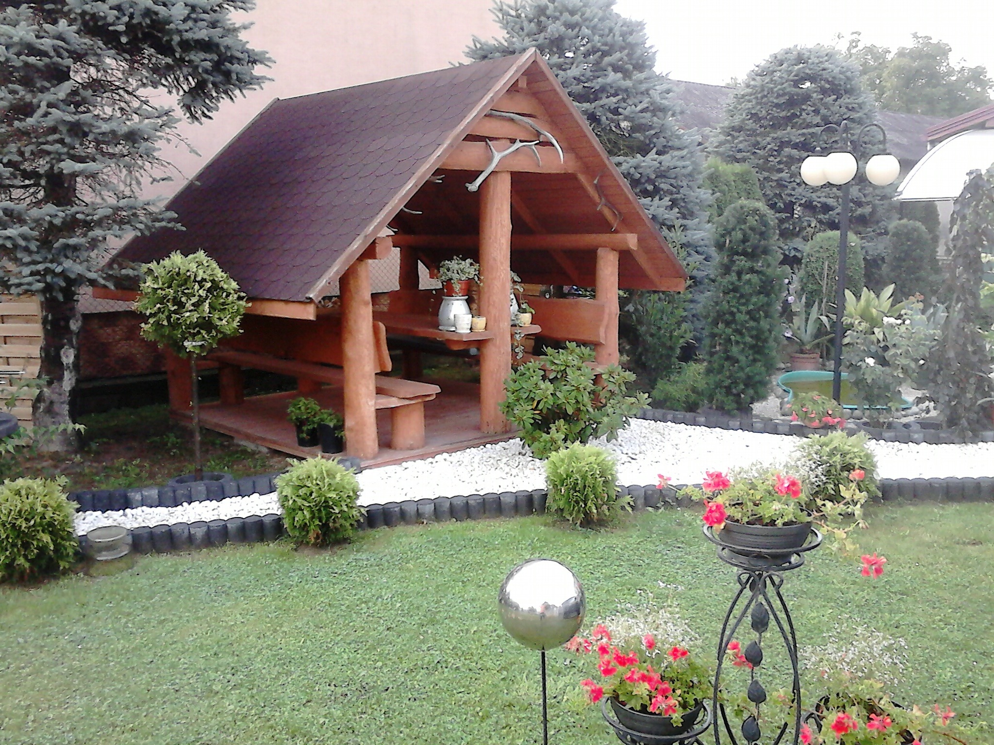 Drewniana altana ogrodowa z brązowym dachem, ozdobiona porożem, otoczona zadbanym trawnikiem, rabatami z kwiatami i dekoracyjnymi kulami